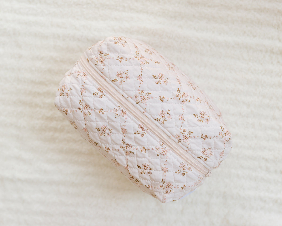 A keepsake / travel bag—a quilted, light pink zippered pouch with a small floral pattern—rests on a soft, cream-colored textured surface, making it an ideal addition to your collection of travel accessories.