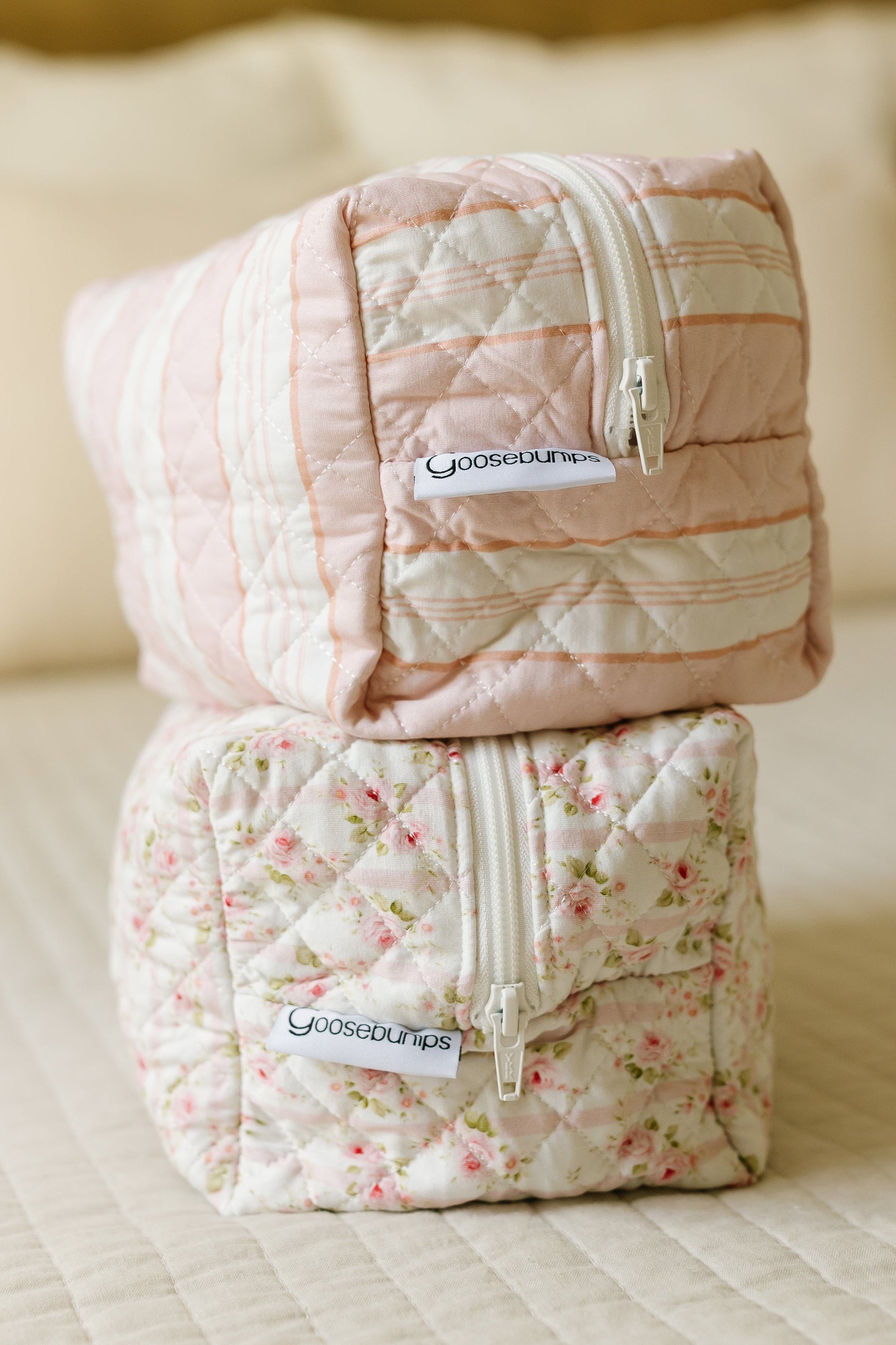 Two “lover girl” travel bags—one pink, one floral—are stacked on a cream bed. Each has a white zipper and Goodsouls label, making them perfect for carrying stylish accessories as Goosebumps Travel Bags.