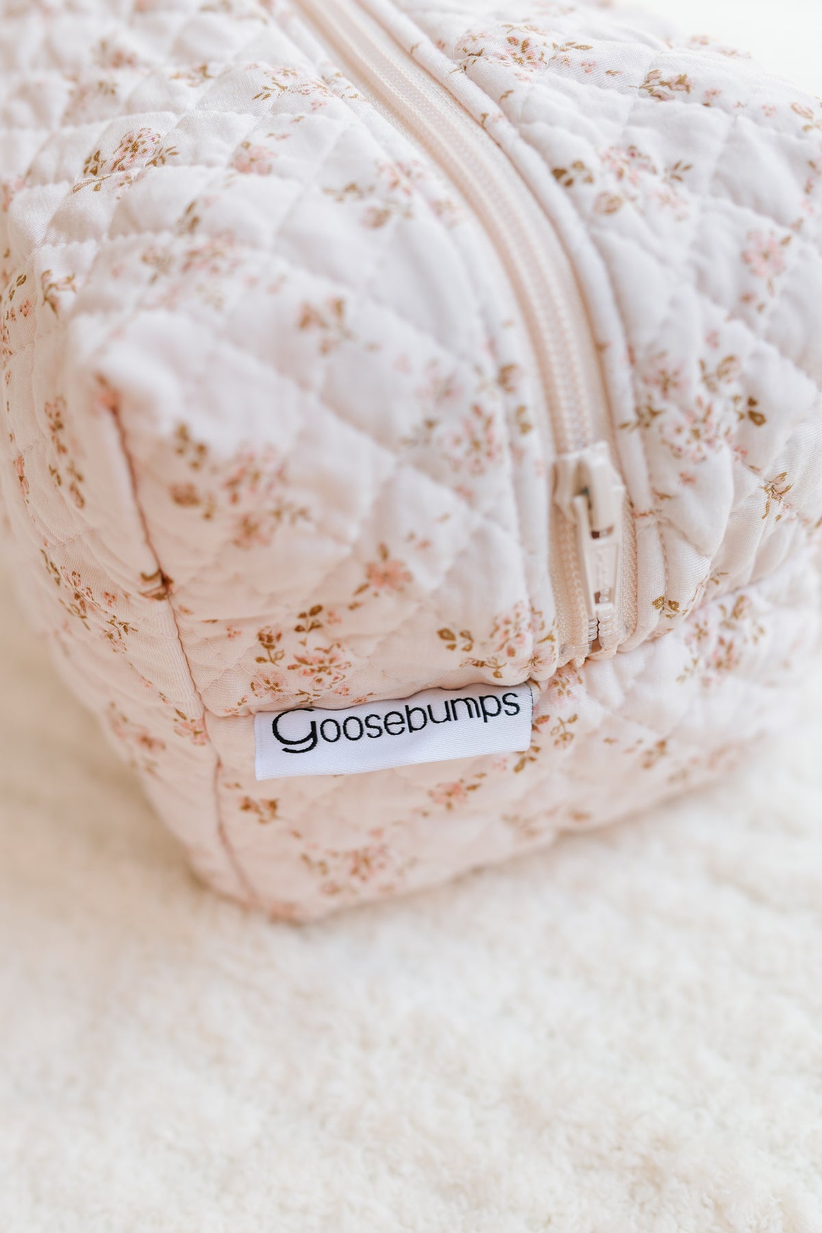 A close-up, high-angle shot showcases a soft, quilted fabric keepsake / travel bag adorned with a light pink floral print. A prominent white fabric tag featuring the Goosebumps brand name in black is attached near the zipper. Perfect for use as a stylish travel accessory or your go-to keepsake / travel bag, this charming pouch rests elegantly on a textured white surface.