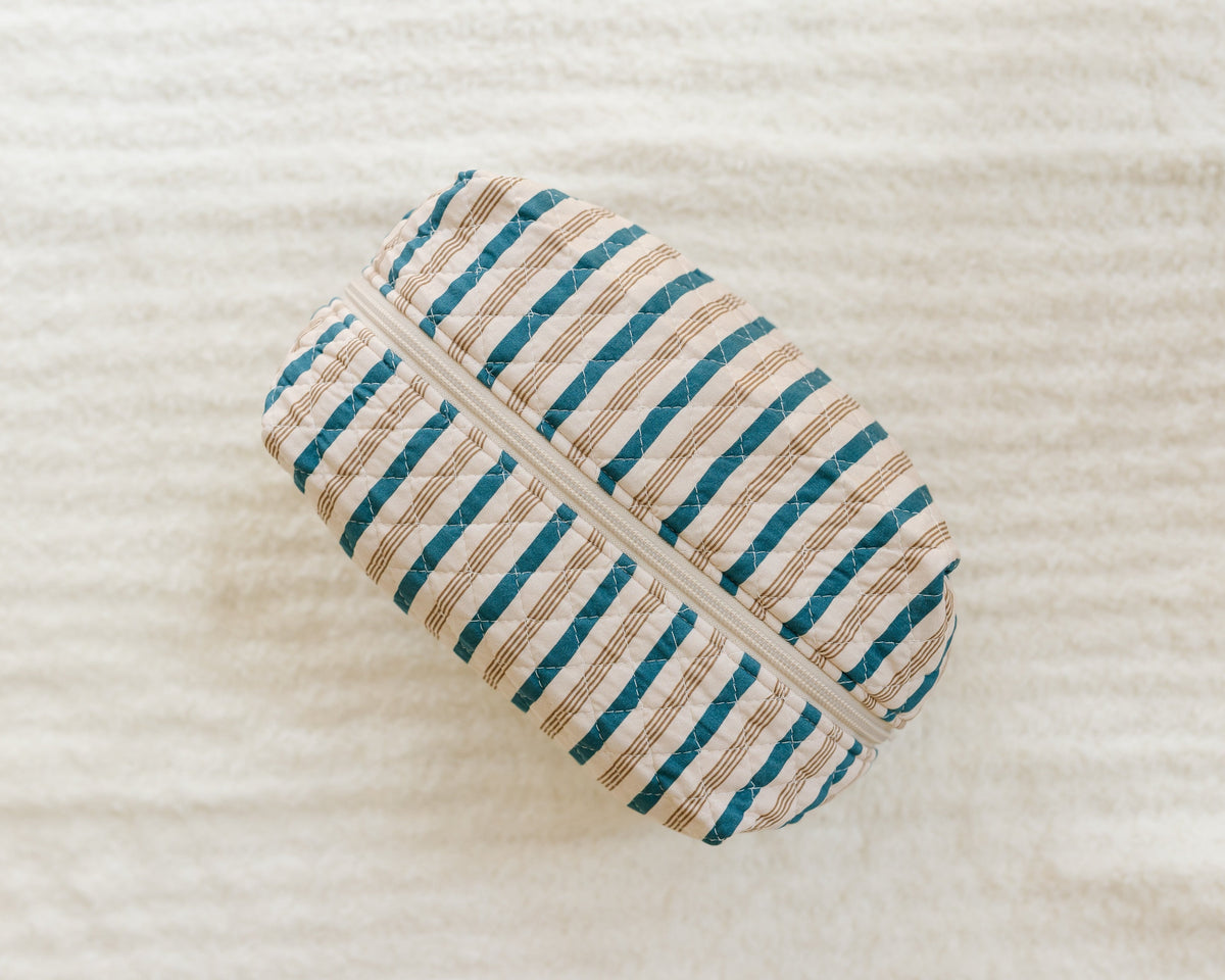 A quilted fabric pouch, named the shoreline / travel bag, with a zipper closure rests on a soft, cream-colored textured surface. Designed for storing travel accessories, this pouch features horizontal blue and beige stripes.