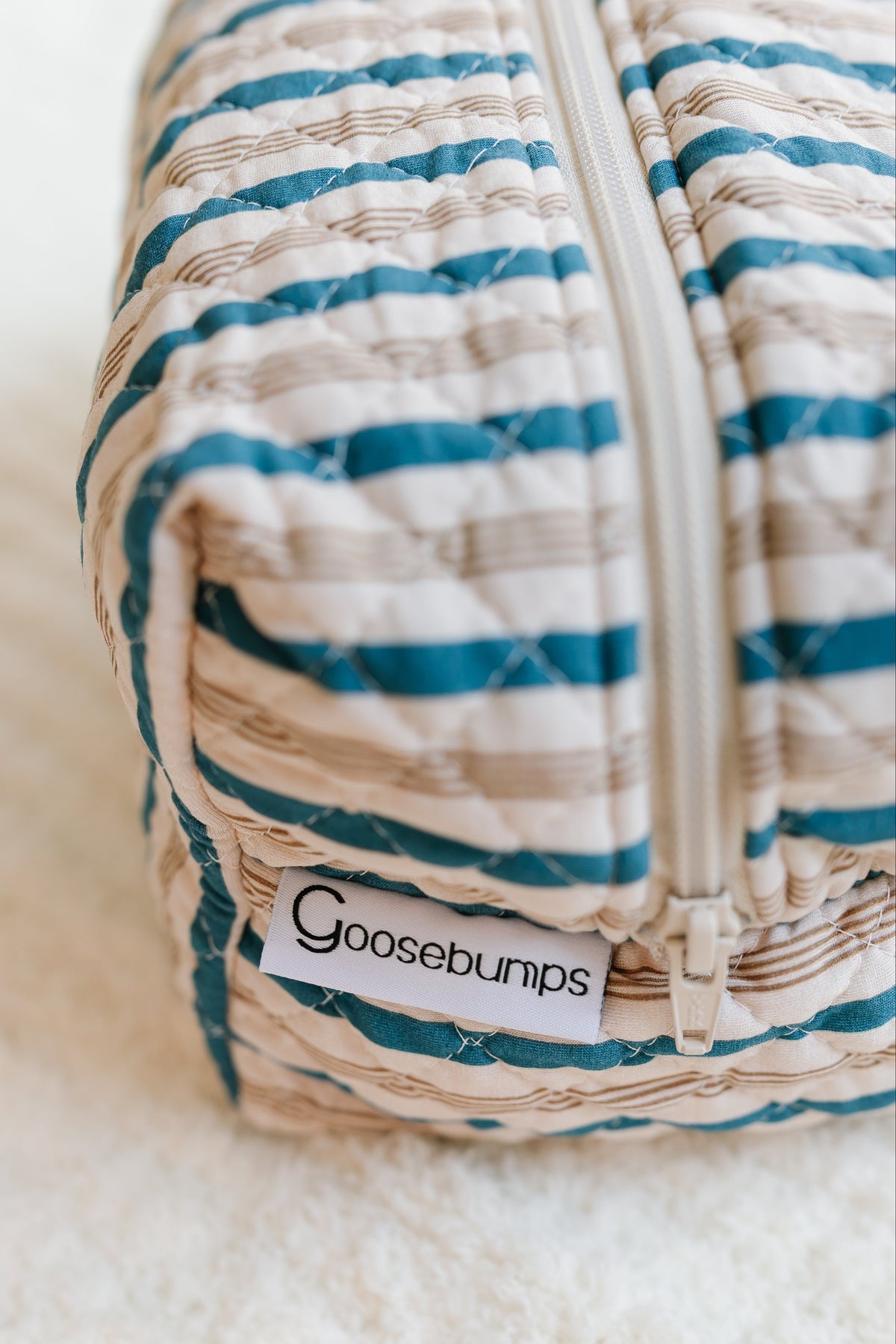 A close-up of the shoreline / travel bag, featuring quilted, striped fabric with a visible zipper and a Goosebumps label, placed on a soft, light-colored surface—perfect for those seeking stylish travel accessories.