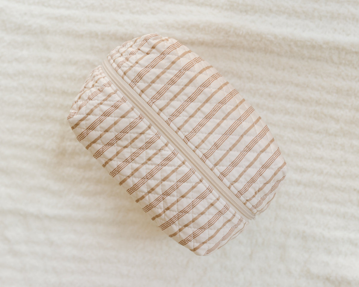 Introducing the toast and honey / travel bag: a rectangular, quilted fabric pouch featuring brown horizontal and vertical stripes with a white zipper, set atop a textured off-white surface—making it an ideal stylish addition to your travel accessories collection or a unique Goosebumps-inspired travel bag.