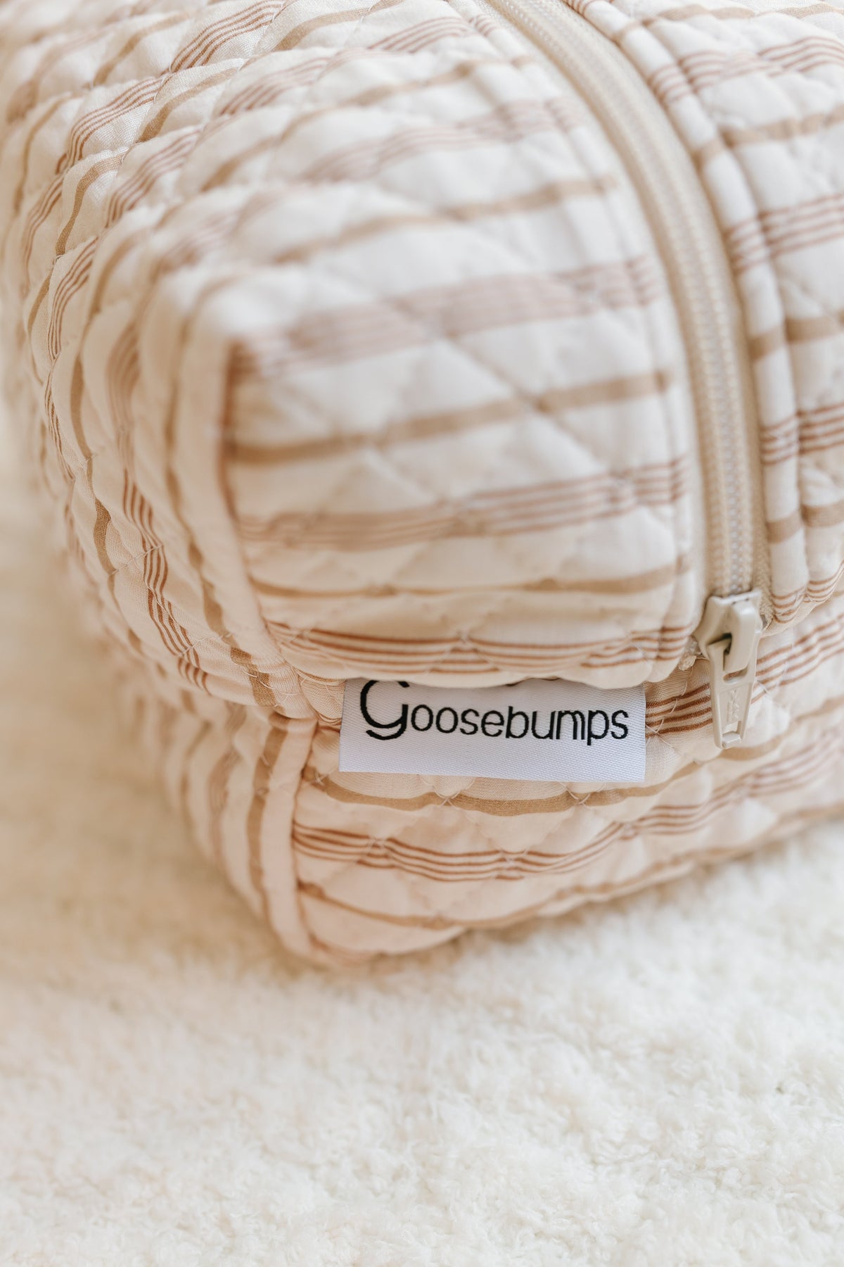 A quilted, beige-striped fabric pouch with a zipper sits on a soft, white surface. The toast and honey / travel bag is perfect for carrying your favorite travel accessories and features a small tag that reads Goosebumps.