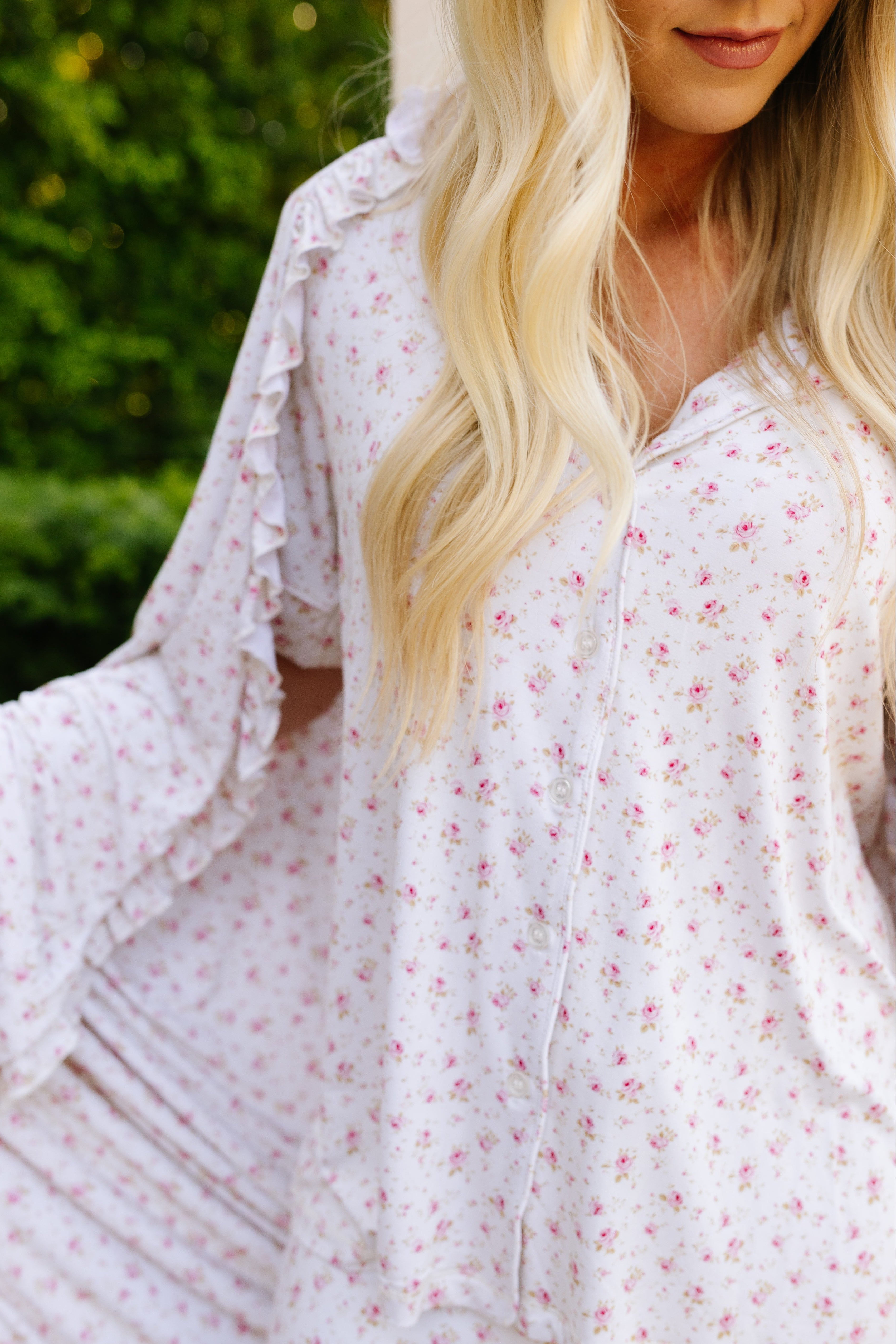 A woman with long blonde hair models the "afternoon tea / ruffle" pajama set, featuring a light floral pattern and ruffled accents. She holds out her robe, while a plush blanket and soft-focus greenery complete the cozy scene.