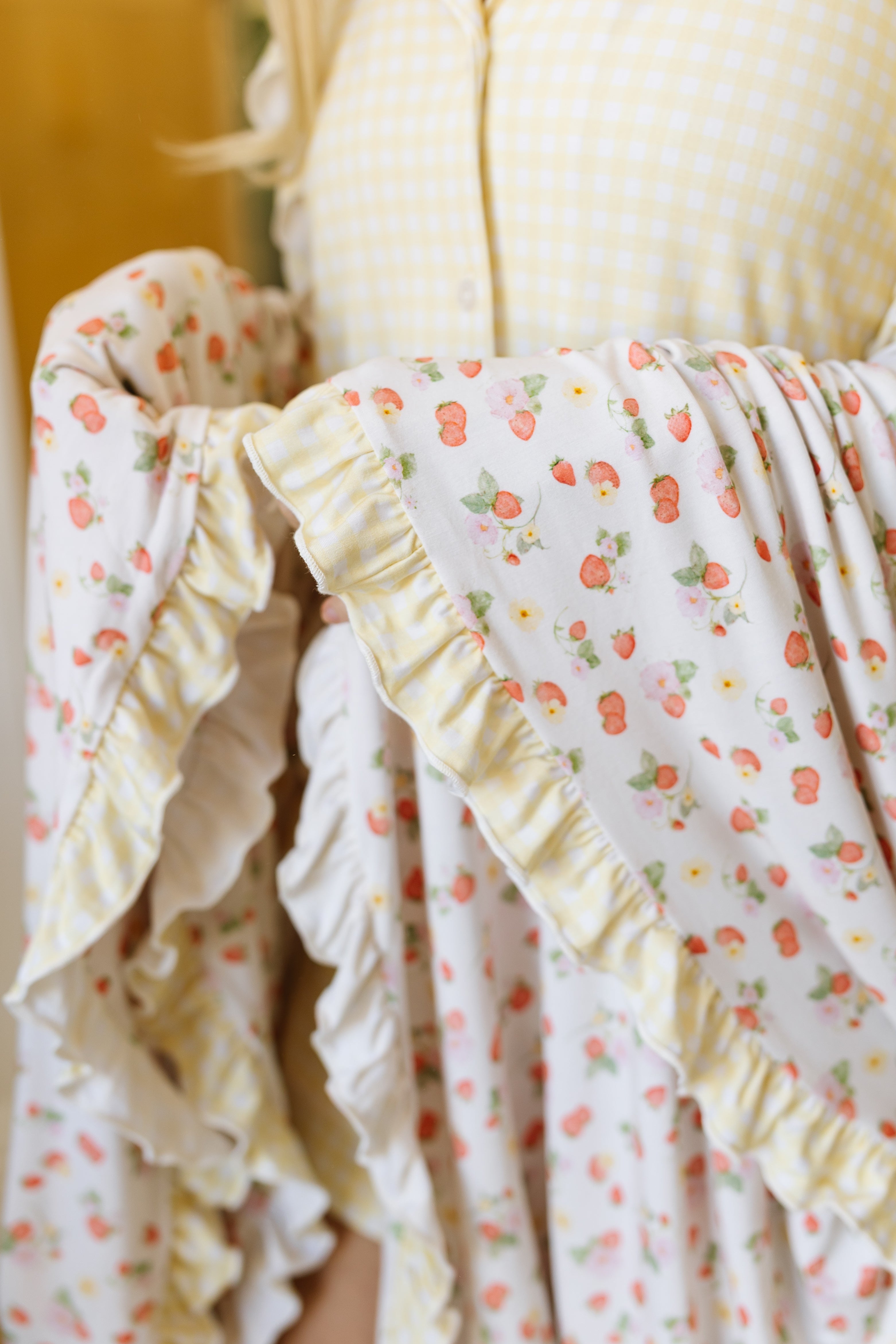 A person in a yellow gingham top holds the "freshly picked w/ sunshine ruffle" travel blanket, featuring strawberry and daisy prints, playful ruffles, and yellow-and-white striped trim. The focus is on its cheerful patterned fabric and details.