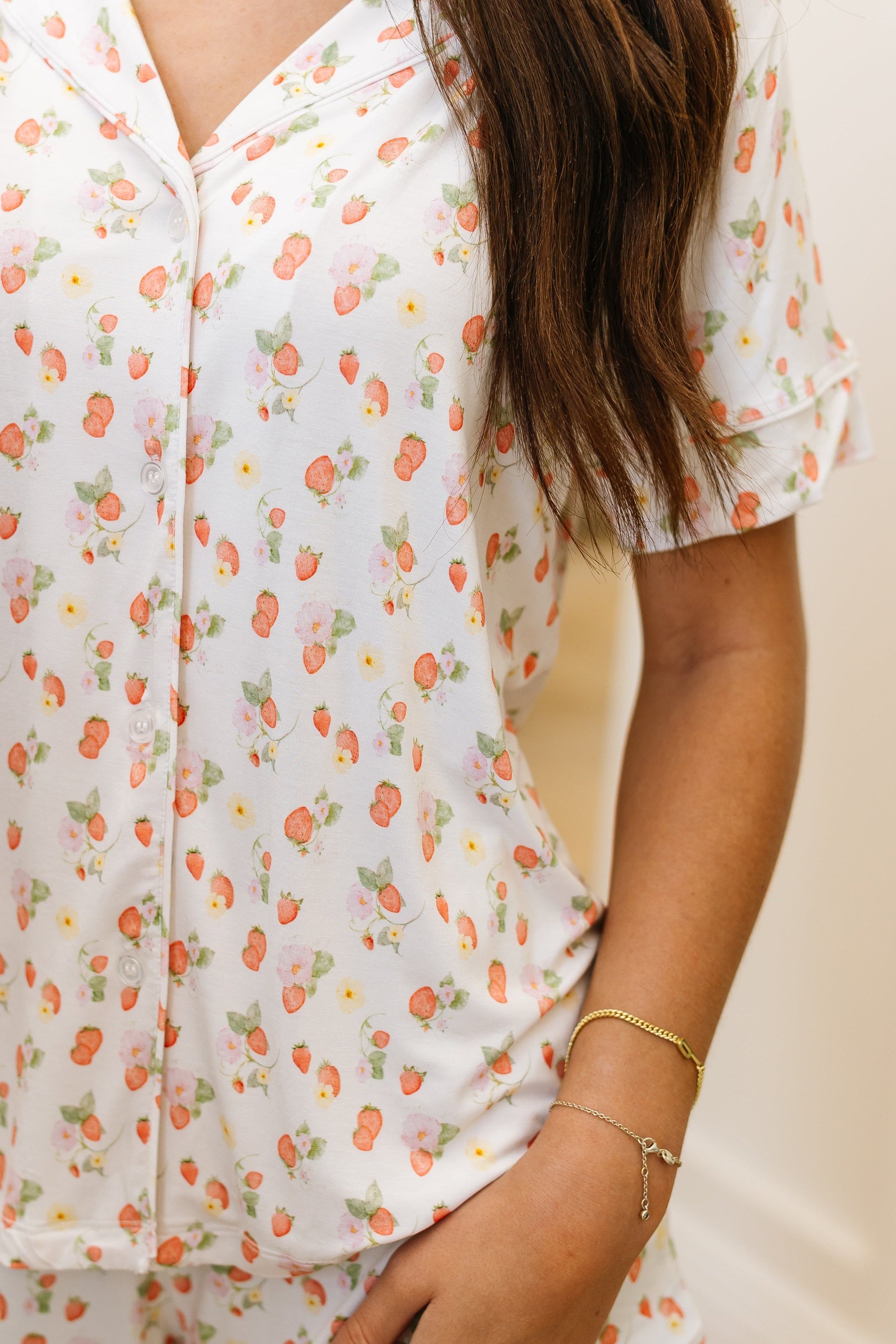 A person wearing the freshly picked pajama set / shorts in white bamboo with a garden strawberry print, featuring short sleeves and buttons. Their long brown hair and a gold bracelet on their wrist are visible.