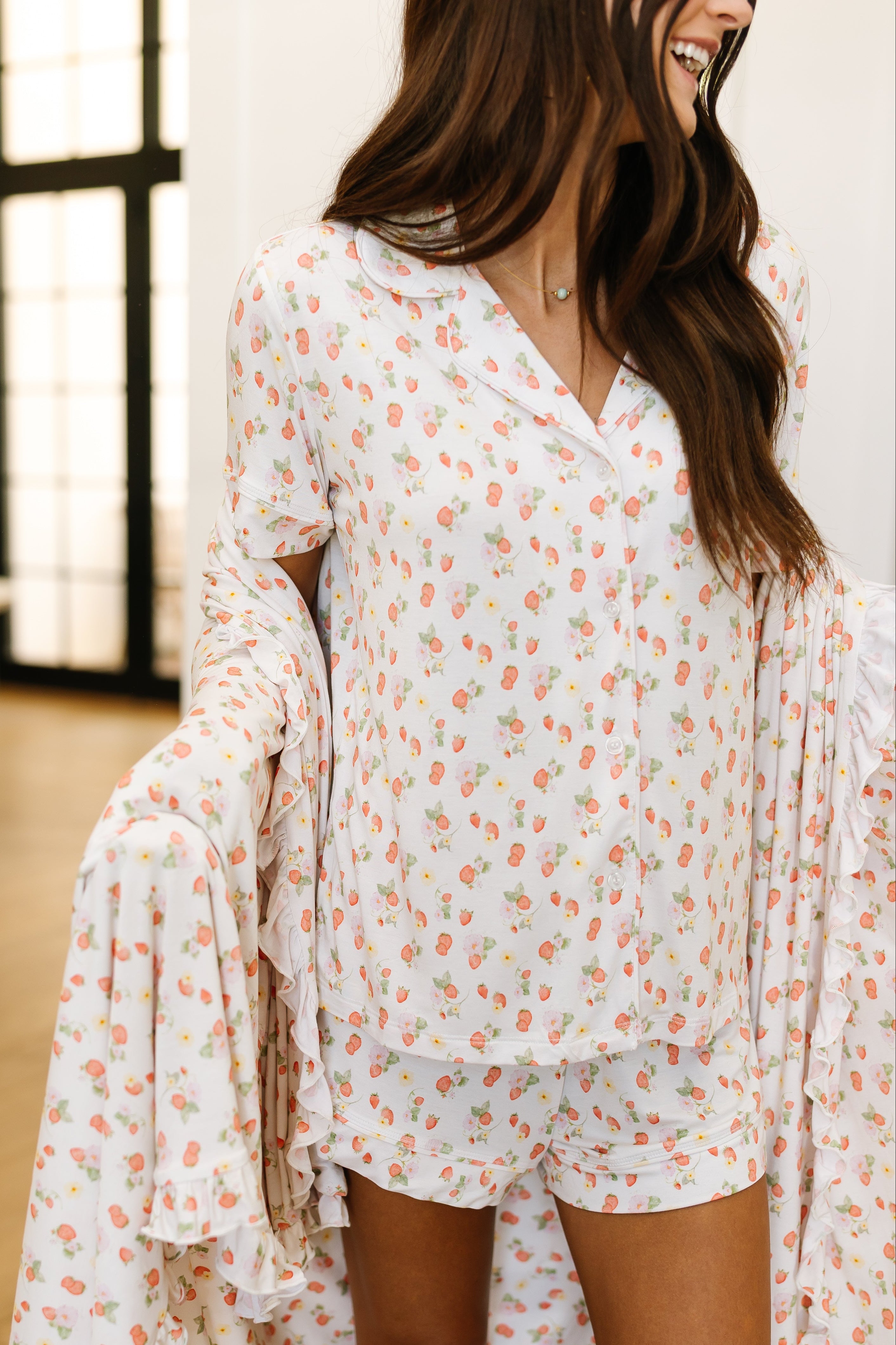 A woman with long brown hair models the freshly picked pajama set with shorts, featuring a pink and orange floral print, indoors by large black-framed windows. She smiles, standing in the matching loungewear.