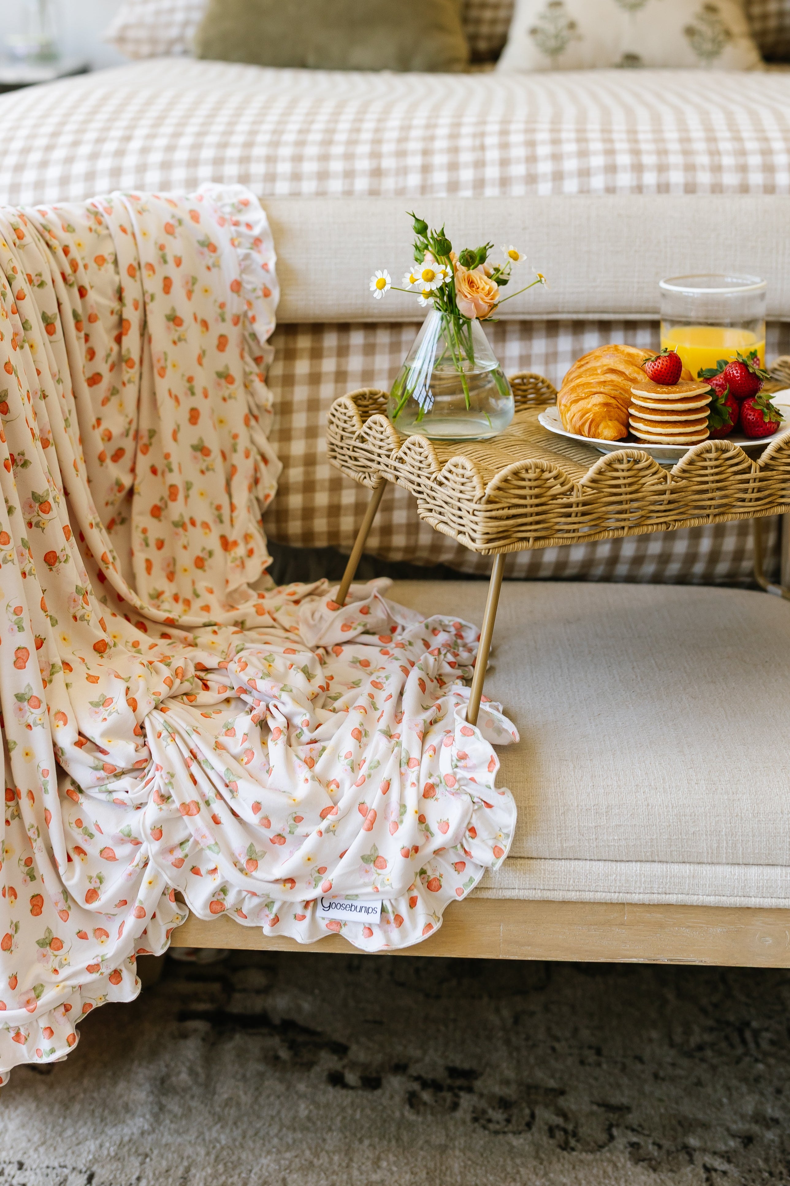 A freshly picked / ruffle blanket drapes over a bench next to a wicker tray with a vase of flowers, croissant, strawberries, orange juice, and pancakes, with a neatly made bed in the background.