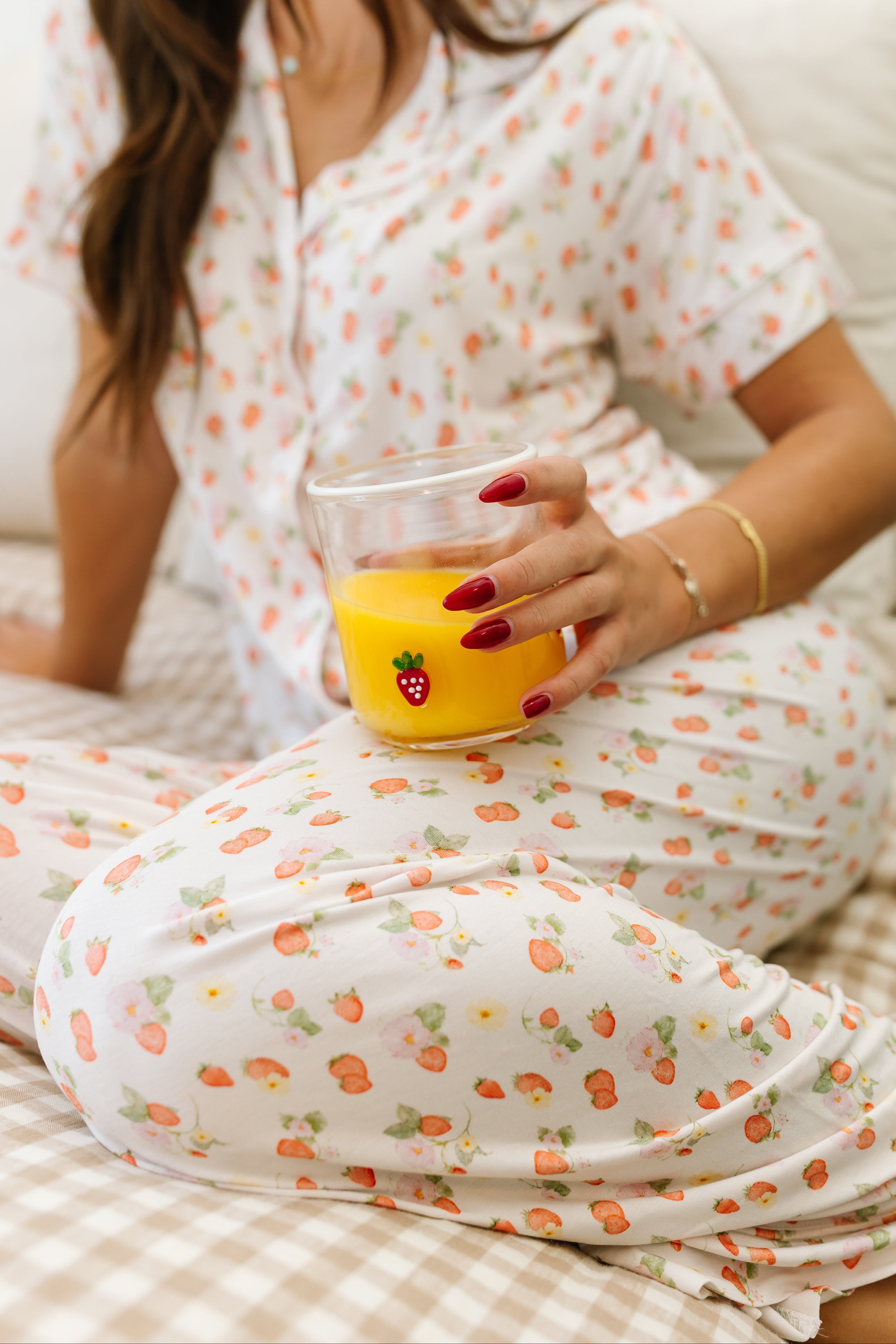 Wearing the freshly picked pajama set / long pant in a garden strawberry print, someone sits on a bed with a glass of orange juice adorned with a strawberry sticker, sporting red nails and a gold bracelet.