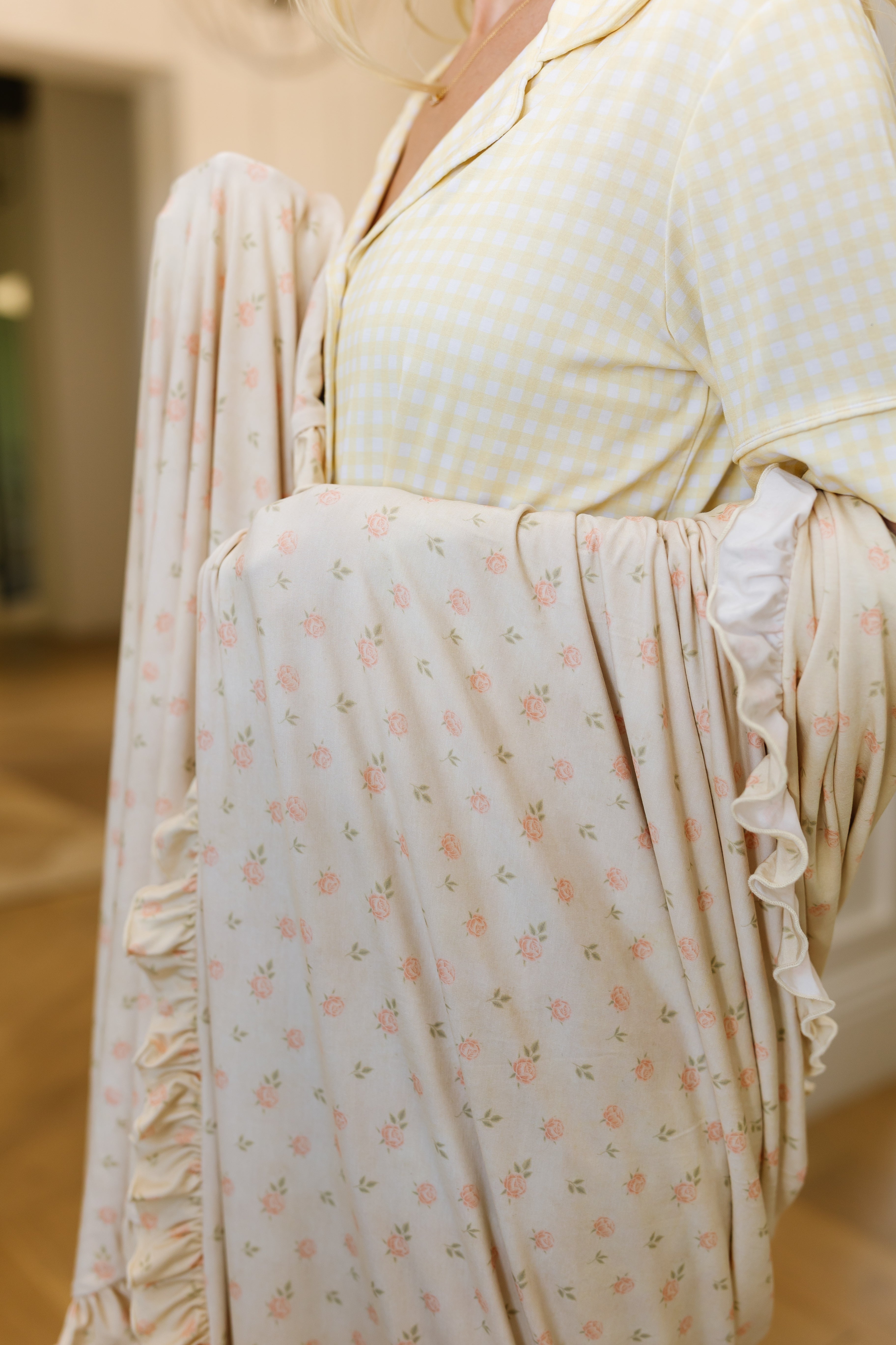 A person in a yellow checkered pajama top holds the "petal / ruffle" blanket, featuring a pink floral pattern and ruffled edge, indoors in soft lighting.
