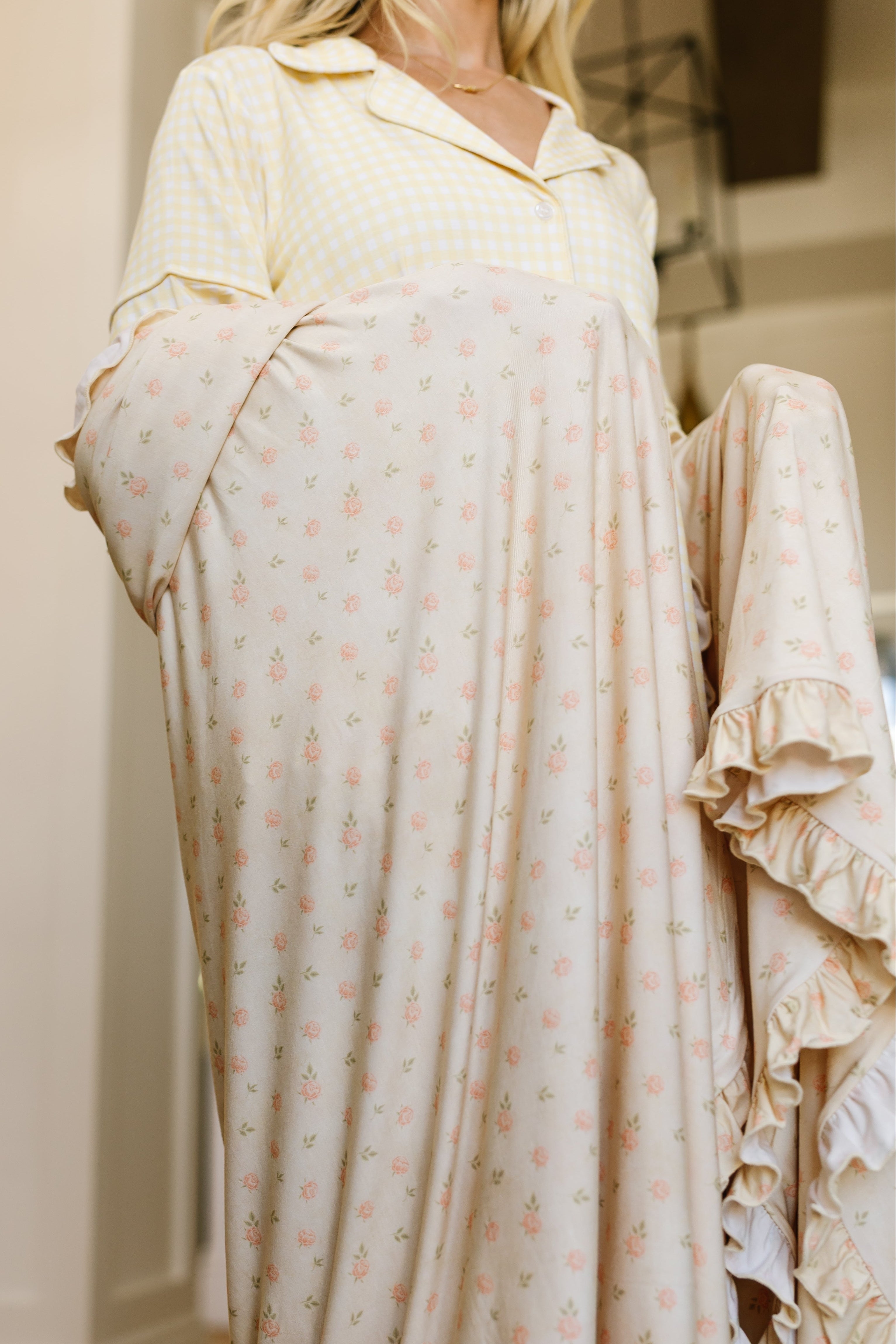 Wearing a yellow and white checkered top, a person holds up the petal / ruffle blanket—a cream-colored, floral-patterned, and ruffled luxurious throw. Their face is out of frame.