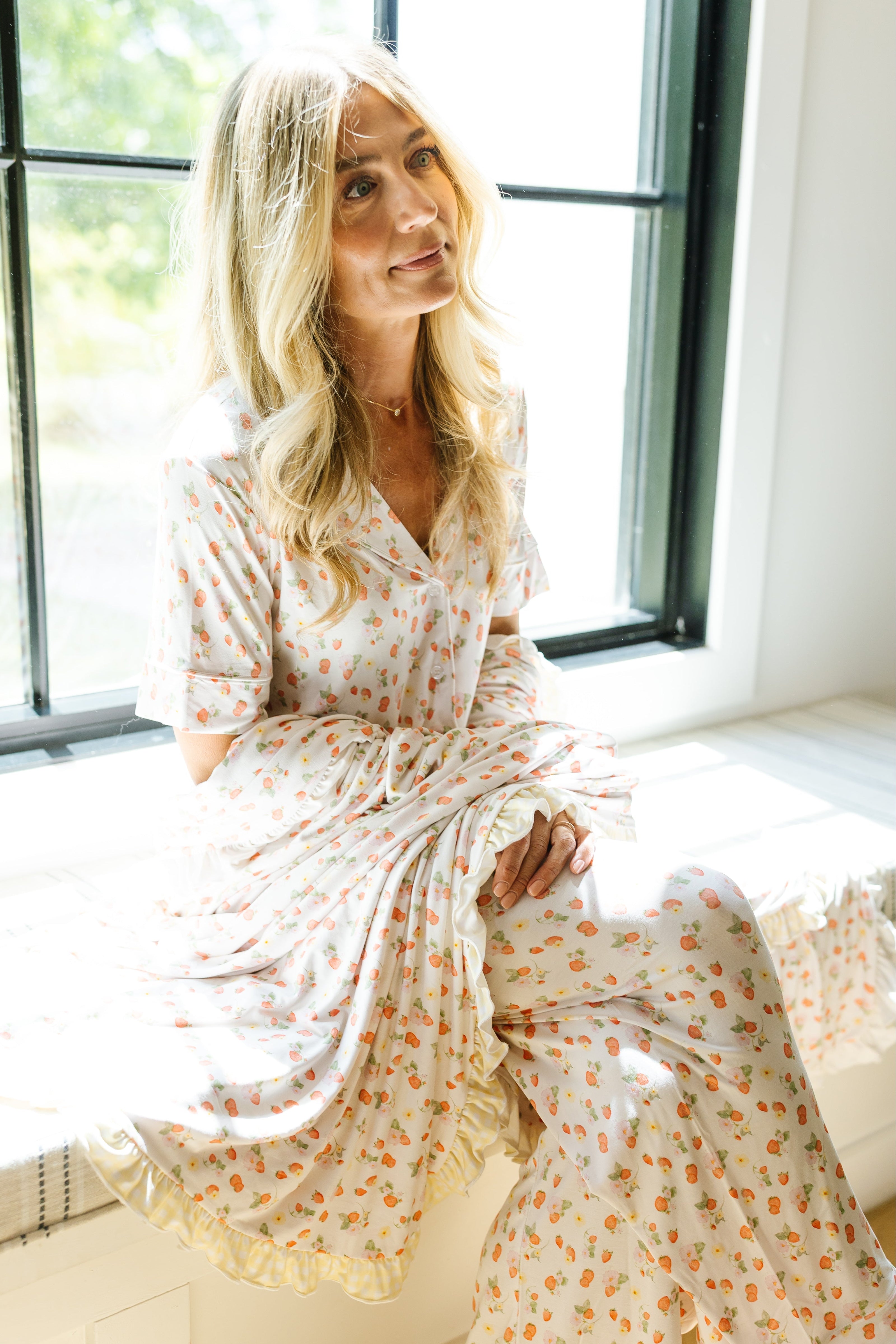 A woman with long blonde hair, wearing the freshly picked pajama set in the garden strawberry print with long pants, sits relaxed on a sunny windowsill, gazing upward—a gentle expression perfect for summer loungewear moments.