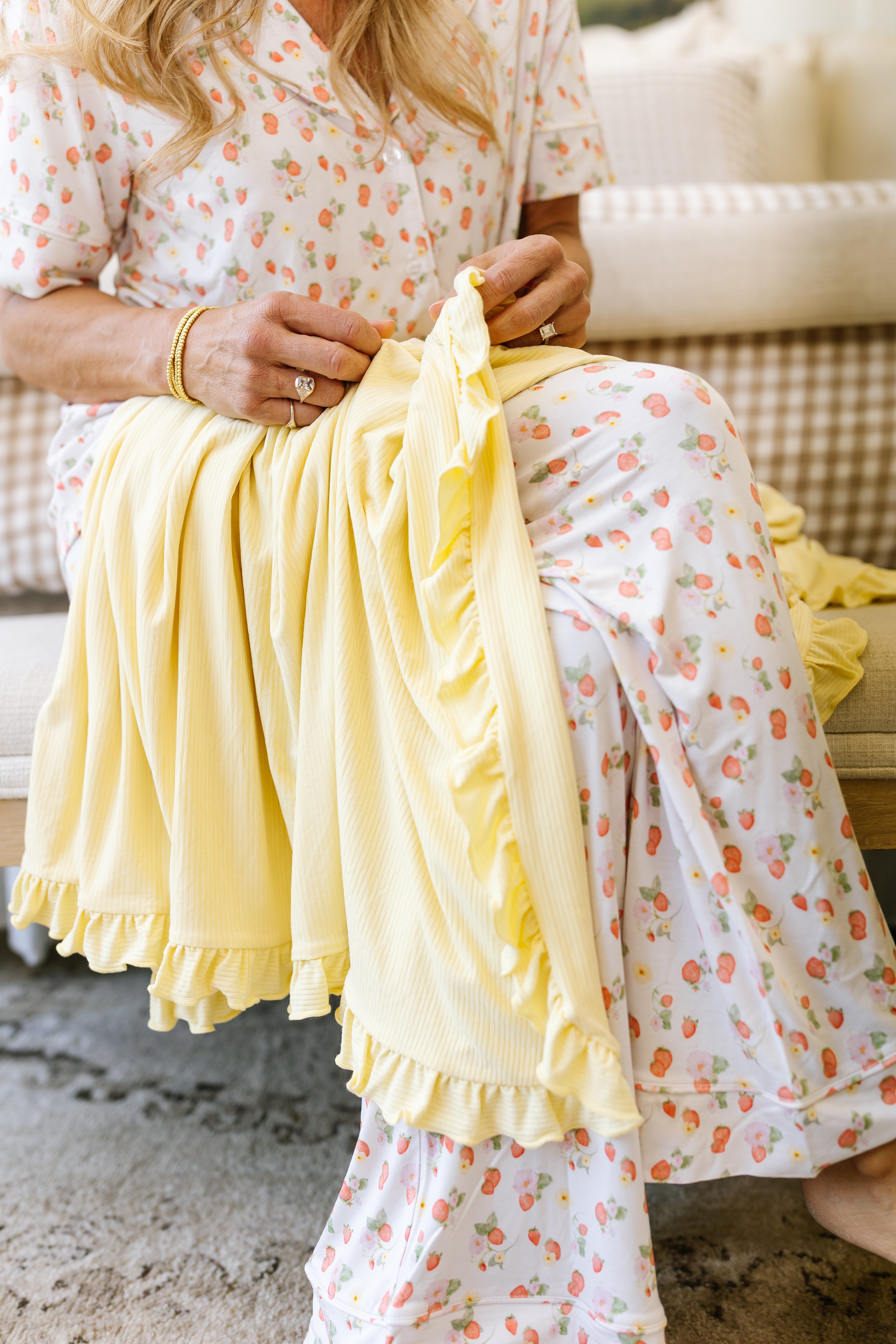 A woman in floral pajamas sits on a cushioned bench, folding the butter / ruffle yellow bamboo blanket in her lap, face out of frame.