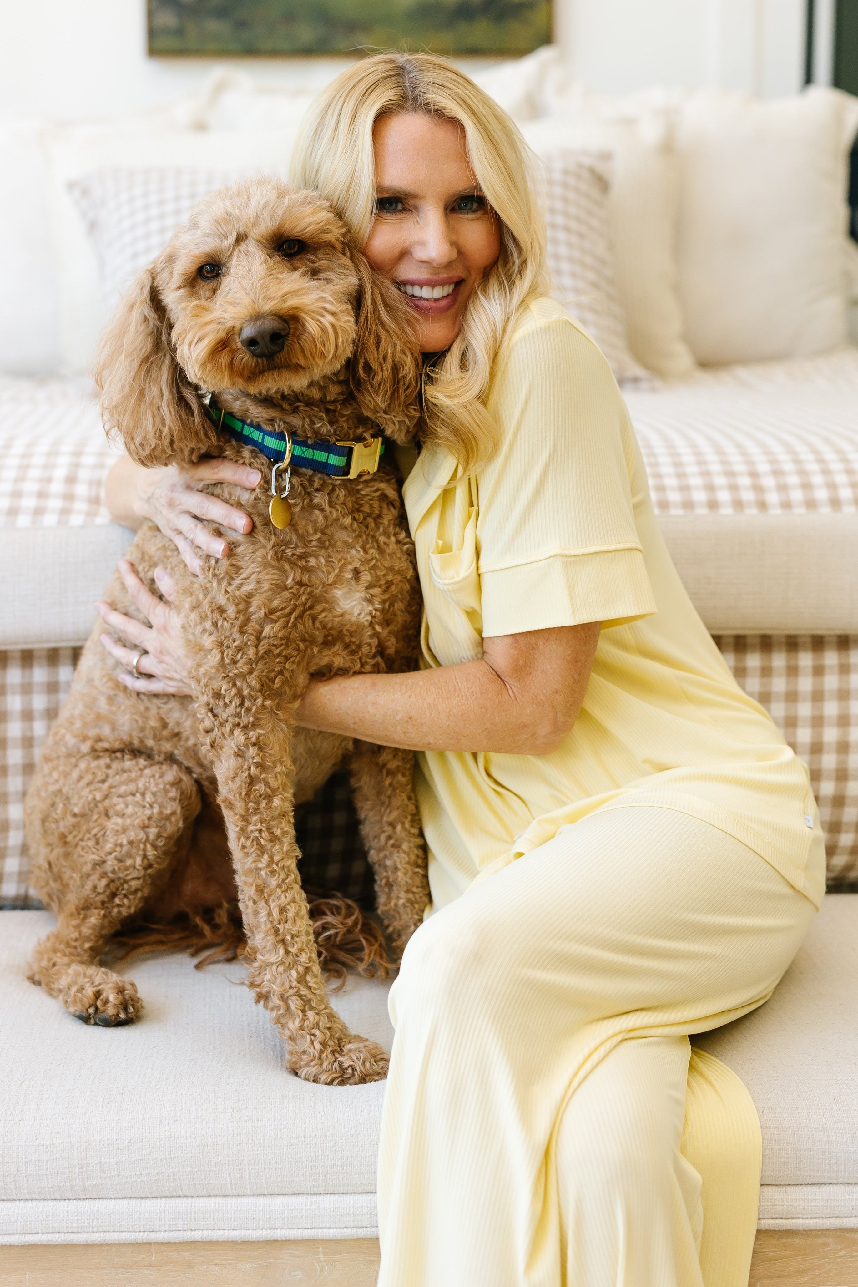 A smiling woman kneels on a couch indoors, hugging a brown, curly-haired dog. She wears the butter short sleeve pajama set; neutral cushions and a checkered pillow are in the background.