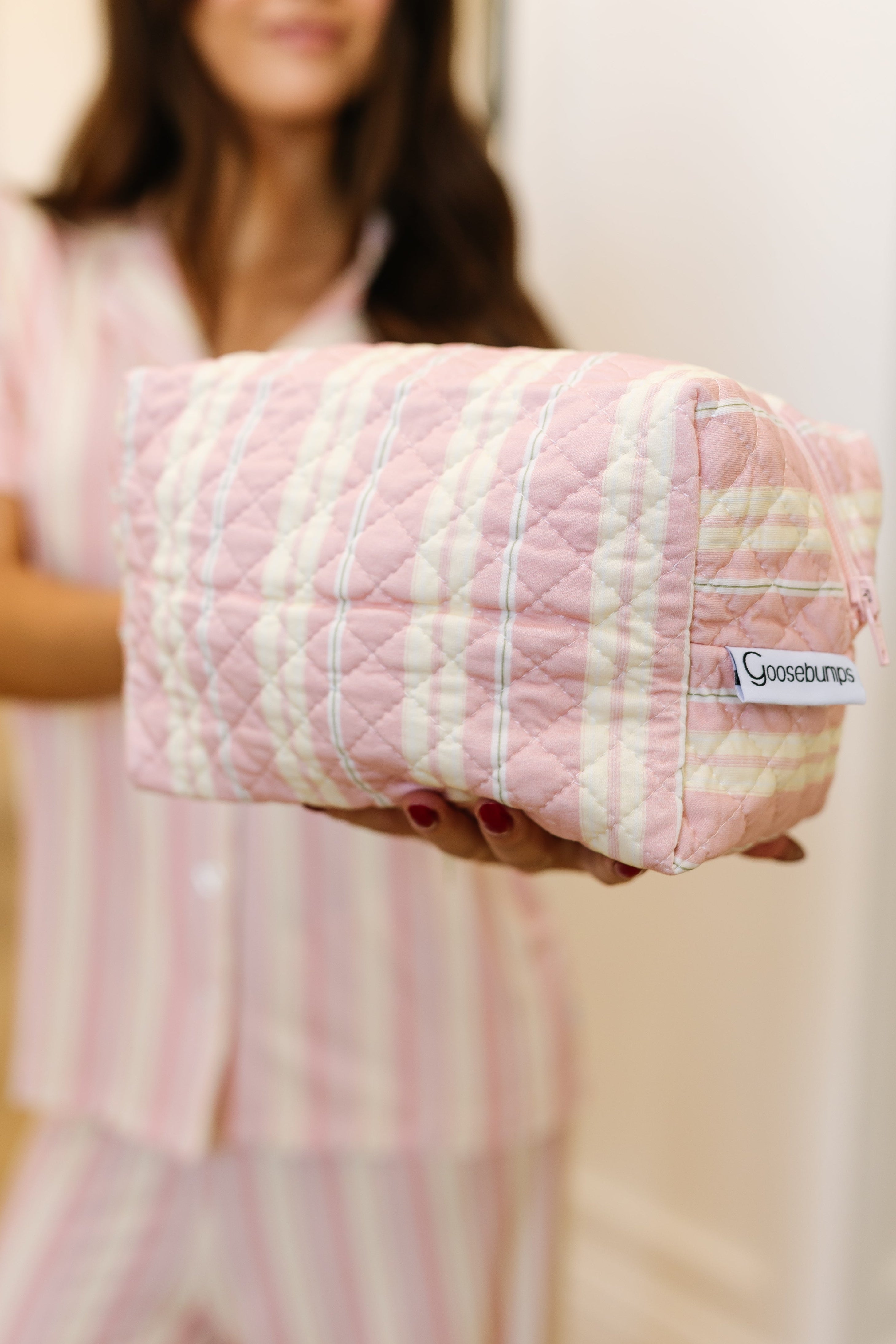 A woman in pink striped pajamas holds out a quilted, pastel pink and yellow plaid sherbet travel bag, perfect for organizing your favorite travel accessories.