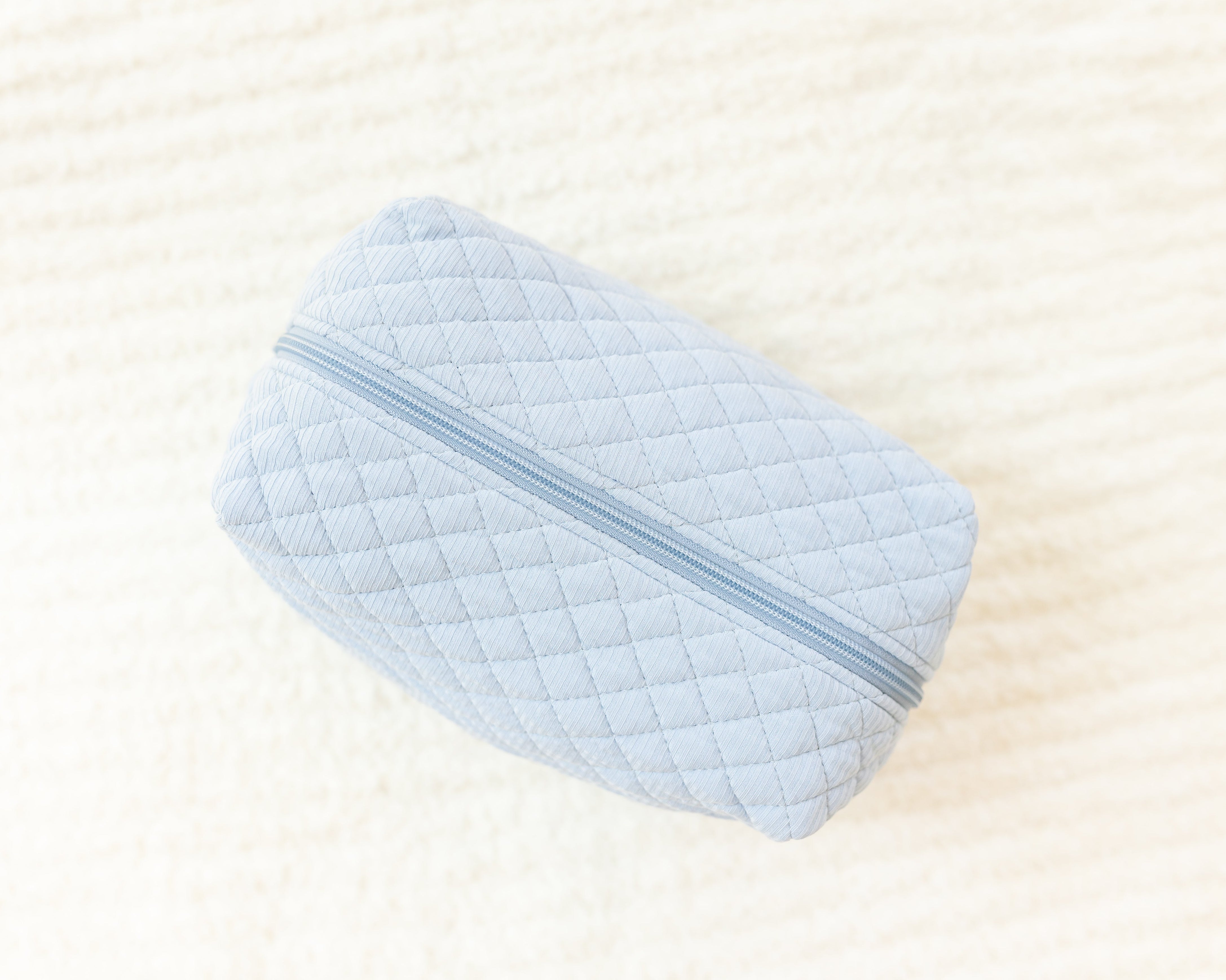 A light blue cove ribbed travel bag with a zipper rests on a soft, cream textured surface—a stylish choice for your travel essentials.