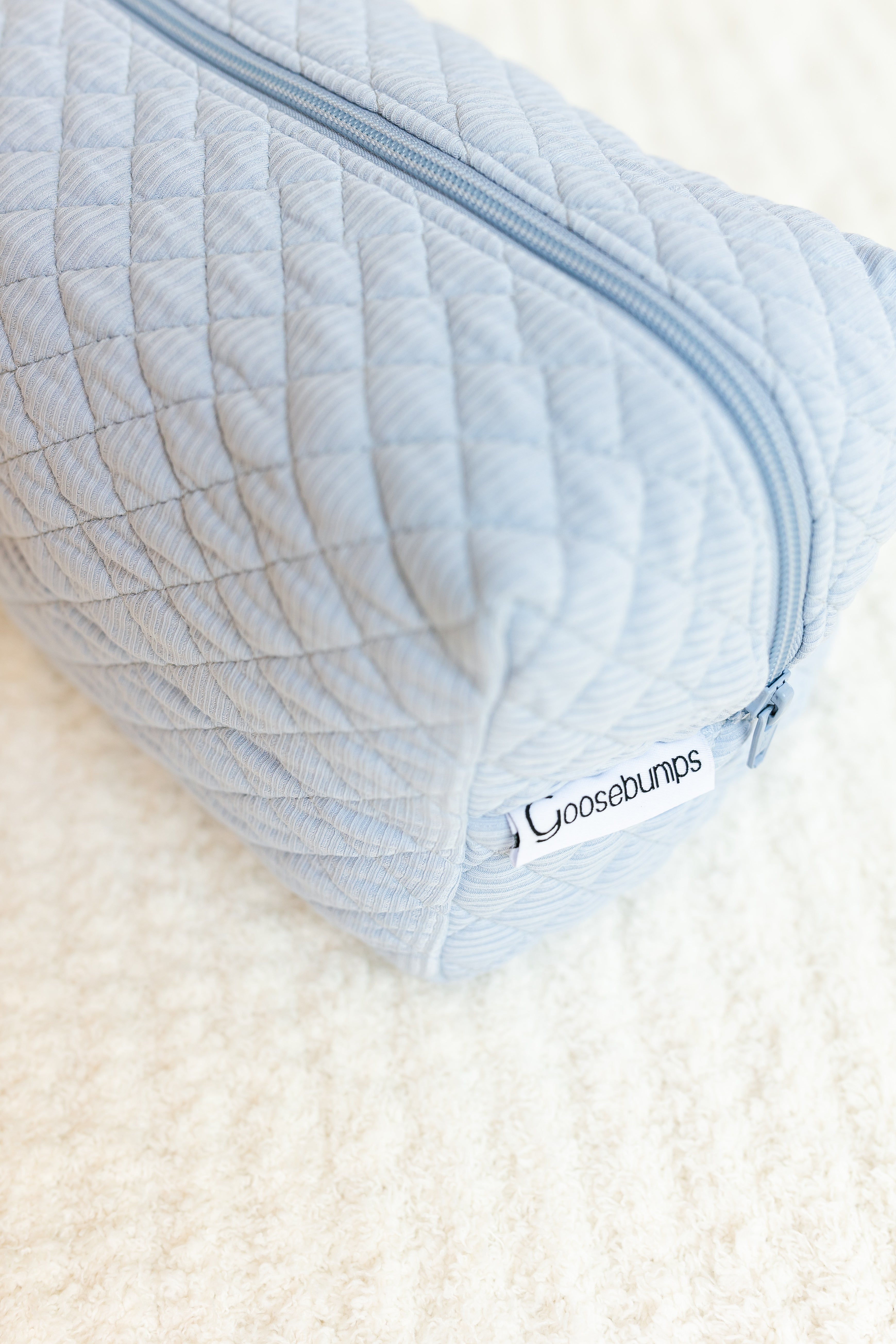 The cove ribbed travel bag in light blue, featuring a zipper and white Goosebumps tag, is perfect for carrying travel accessories and is shown resting on a soft white textured surface.