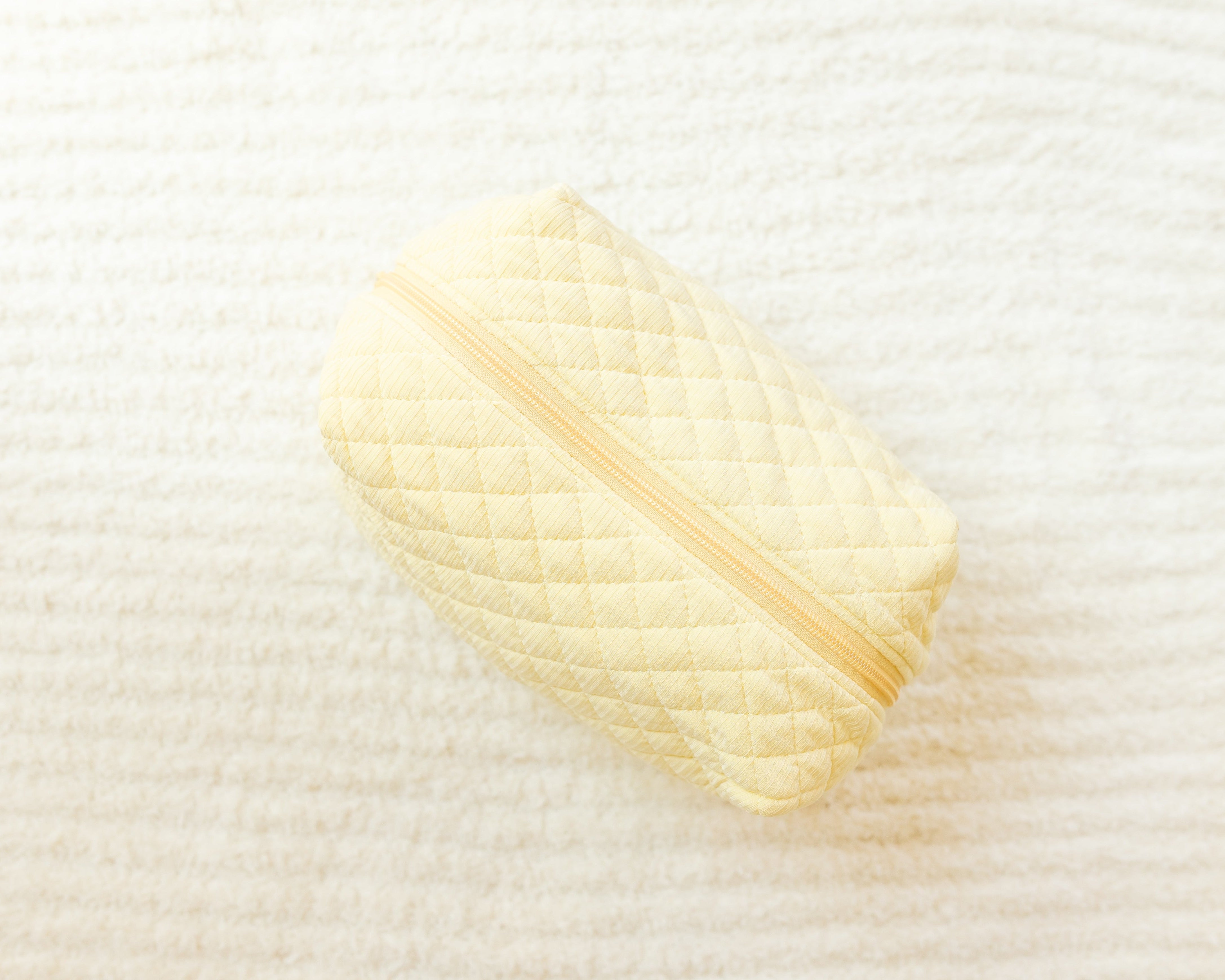 The butter / travel bag is a small, quilted light yellow organizer, ideal for travel accessories, resting on a textured off-white surface.