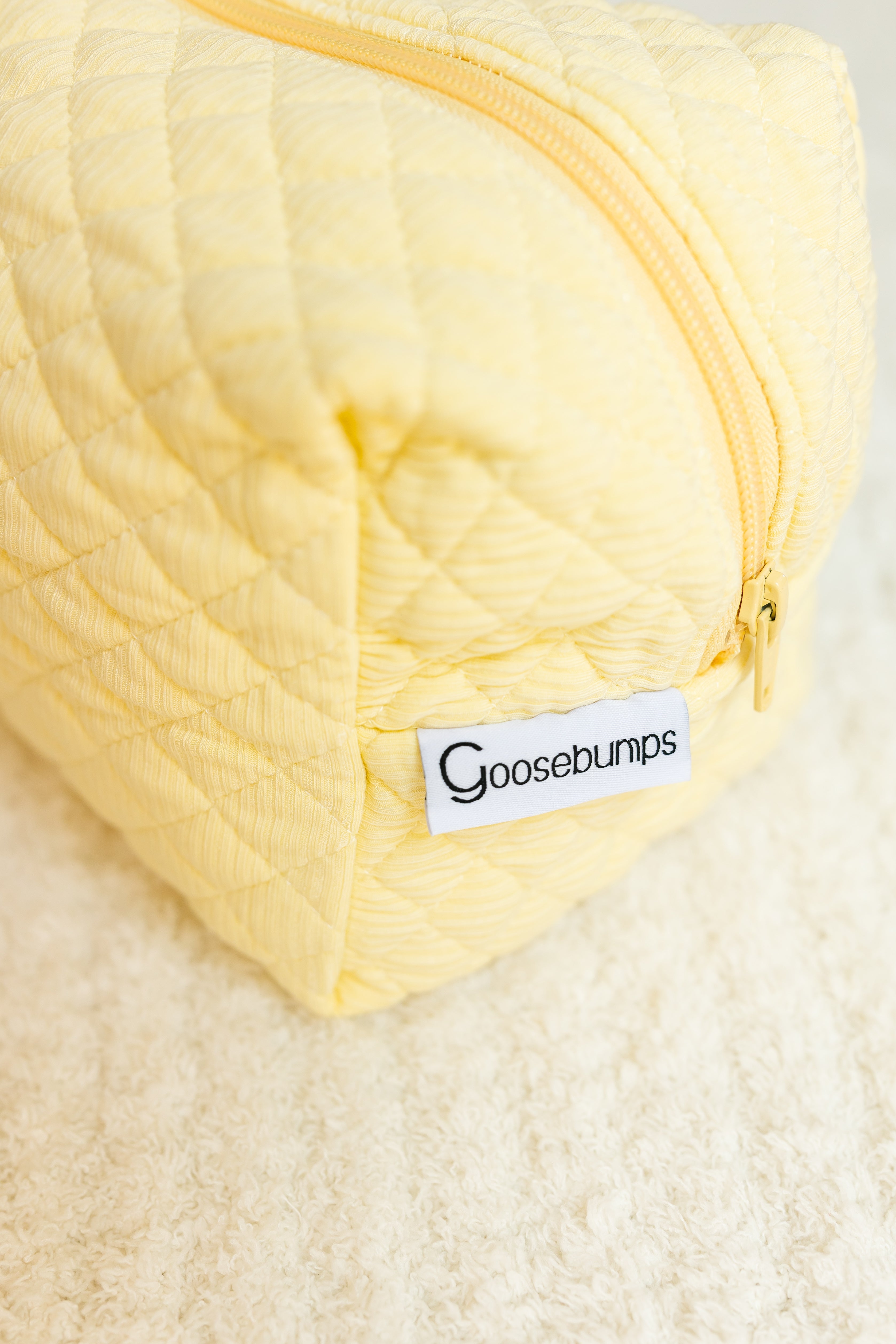 A close-up of the butter / travel bag, a yellow quilted bag with zipper and white label, ideal for storing travel accessories, shown resting on a white textured surface.