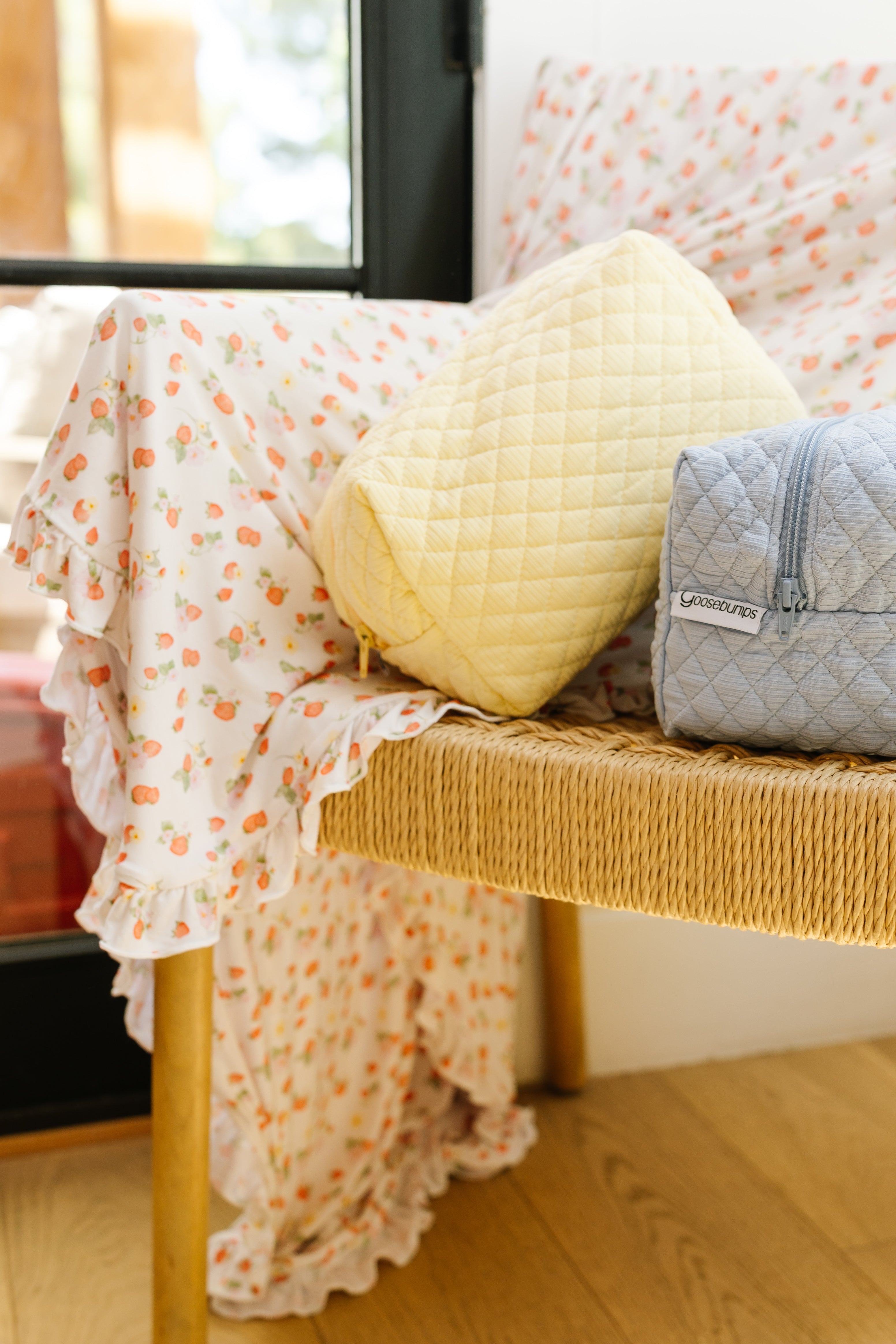 A butter/travel bag sits by a sunlit window alongside a yellow quilted pouch and a floral ruffled blanket draped over a wooden woven bench—ideal for organizing your travel essentials.