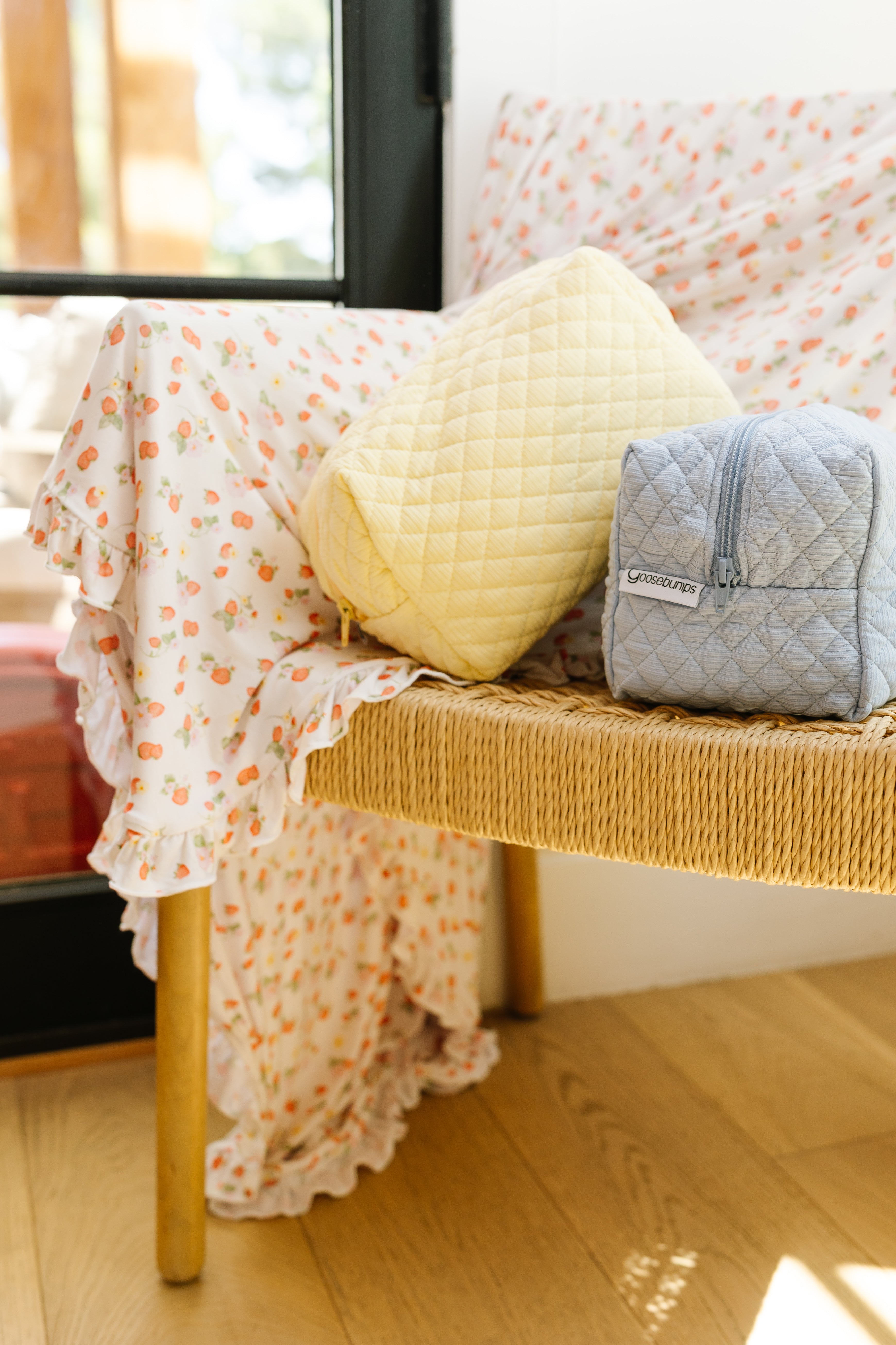 A woven bench with a floral-patterned blanket holds a yellow quilted pouch and a light blue cove ribbed travel bag, perfect for organizing accessories. The bench rests on a wooden floor beside a glass door.