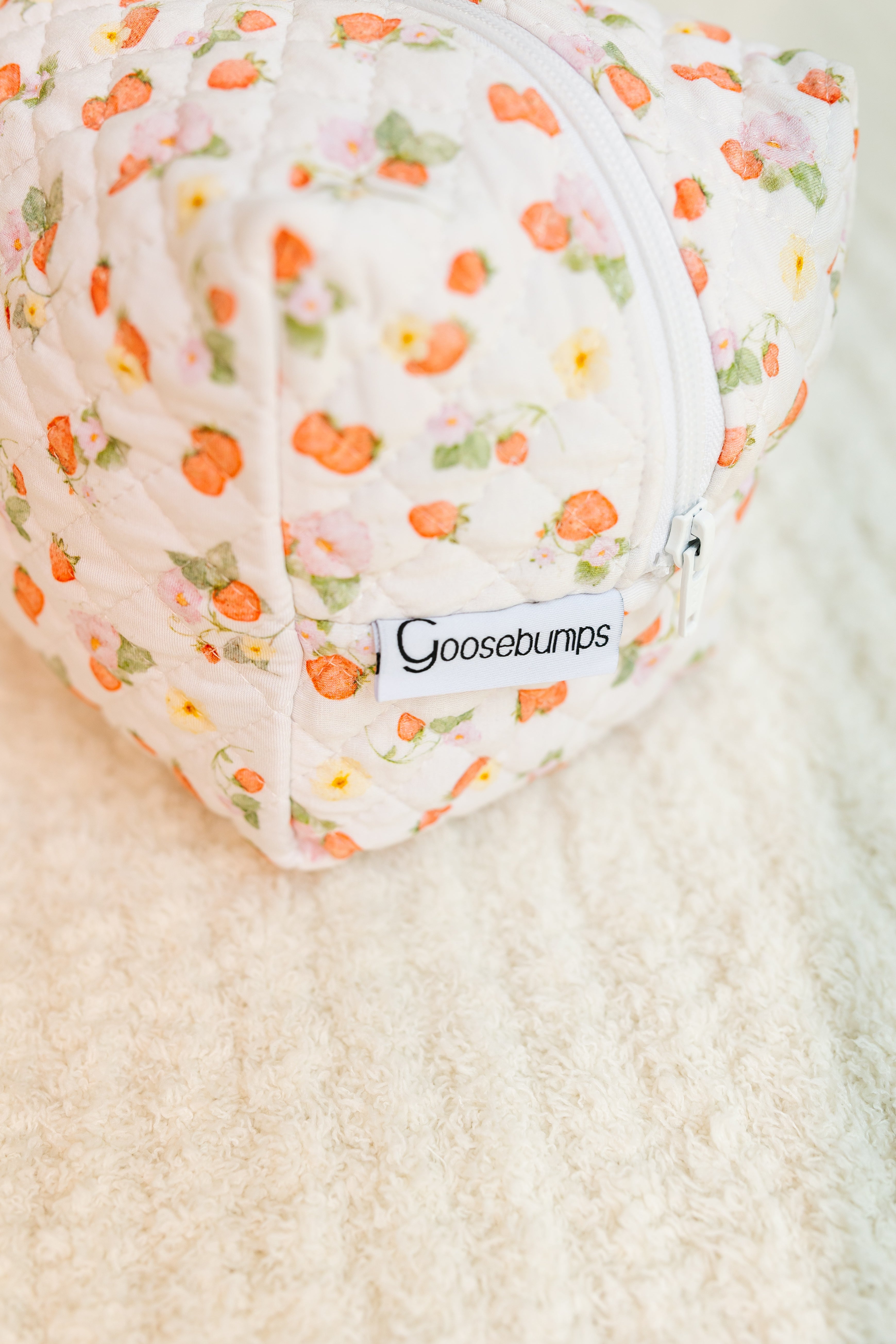 A quilted fabric pouch with a floral pattern, white zipper, and stitched "freshly picked / travel bag" label rests on a textured cream surface—an ideal travel bag for stylish organization.
