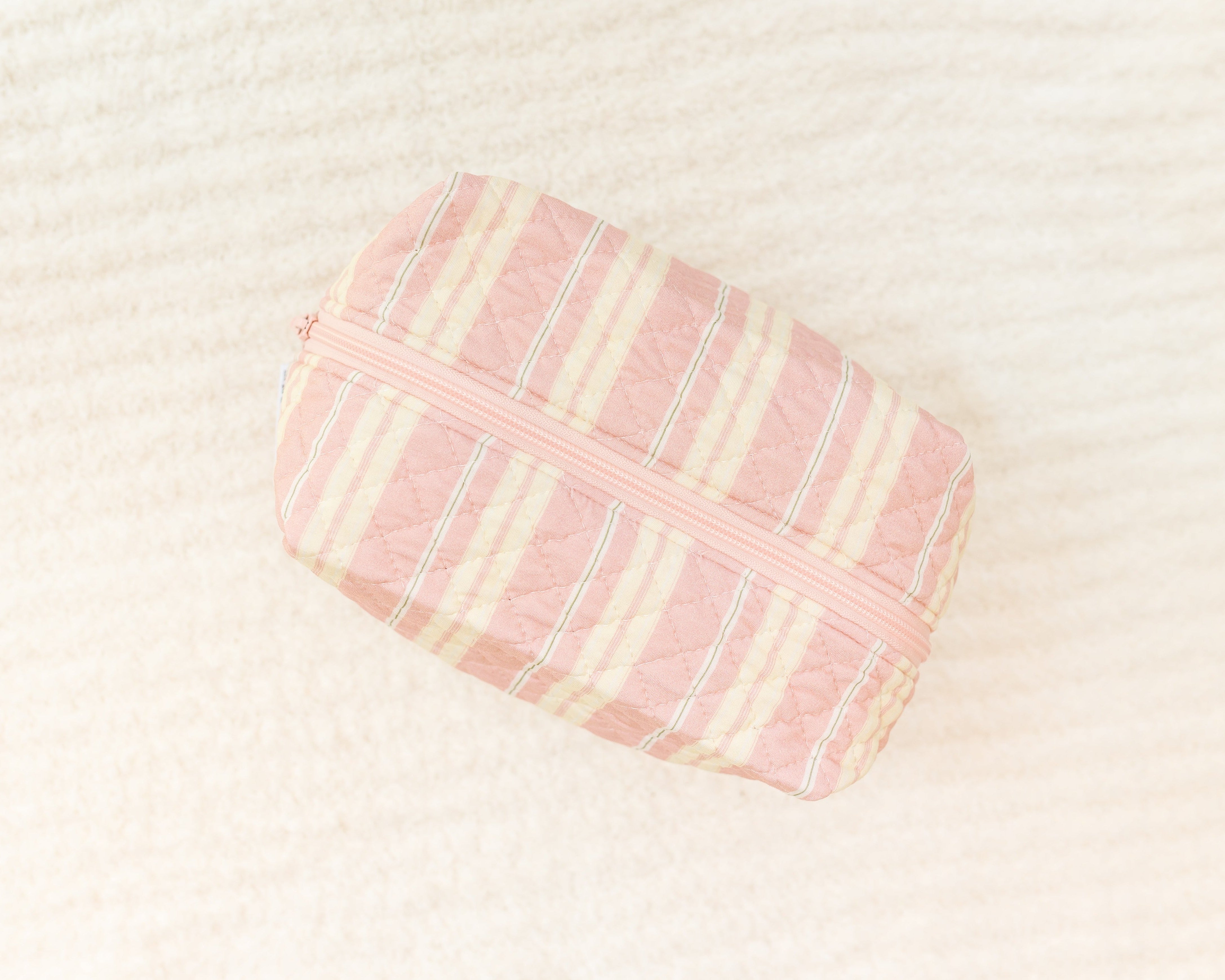 The sherbet travel bag is a small, quilted bag with pink and cream stripes and a pink zipper, shown on a soft off-white surface—ideal for organizing your travel accessories in style.