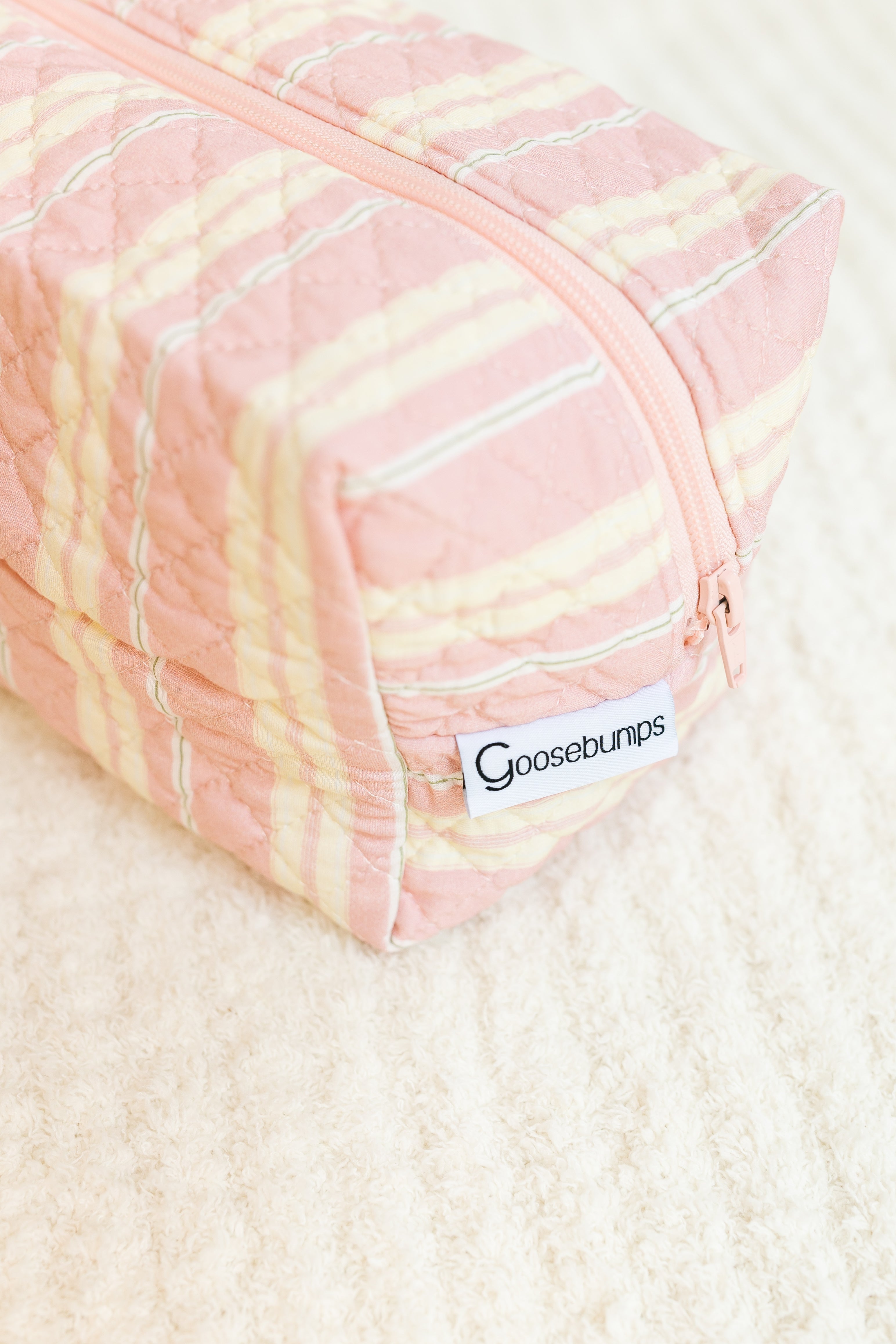 The sherbet travel bag is a soft pink quilted pouch with a peach zipper and yellow plaid pattern. Featuring a “Goosebumps” sewn label, it's perfect for travel accessories and is pictured resting on a textured cream surface.