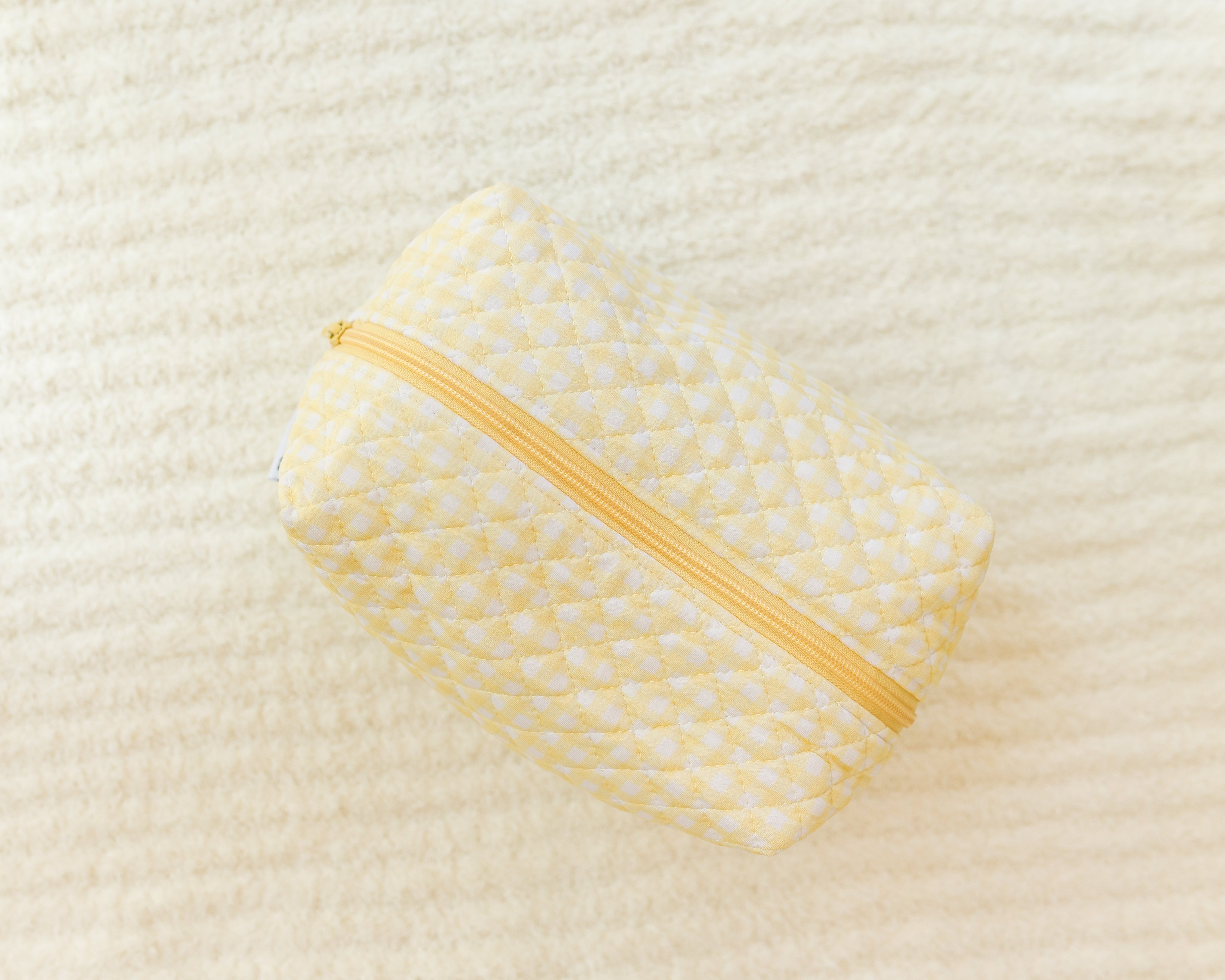 The sunshine / travel bag is a yellow quilted pouch with a yellow zipper and subtle white polka dots, ideal for carrying travel essentials. It’s shown on a soft, cream-colored textured surface.