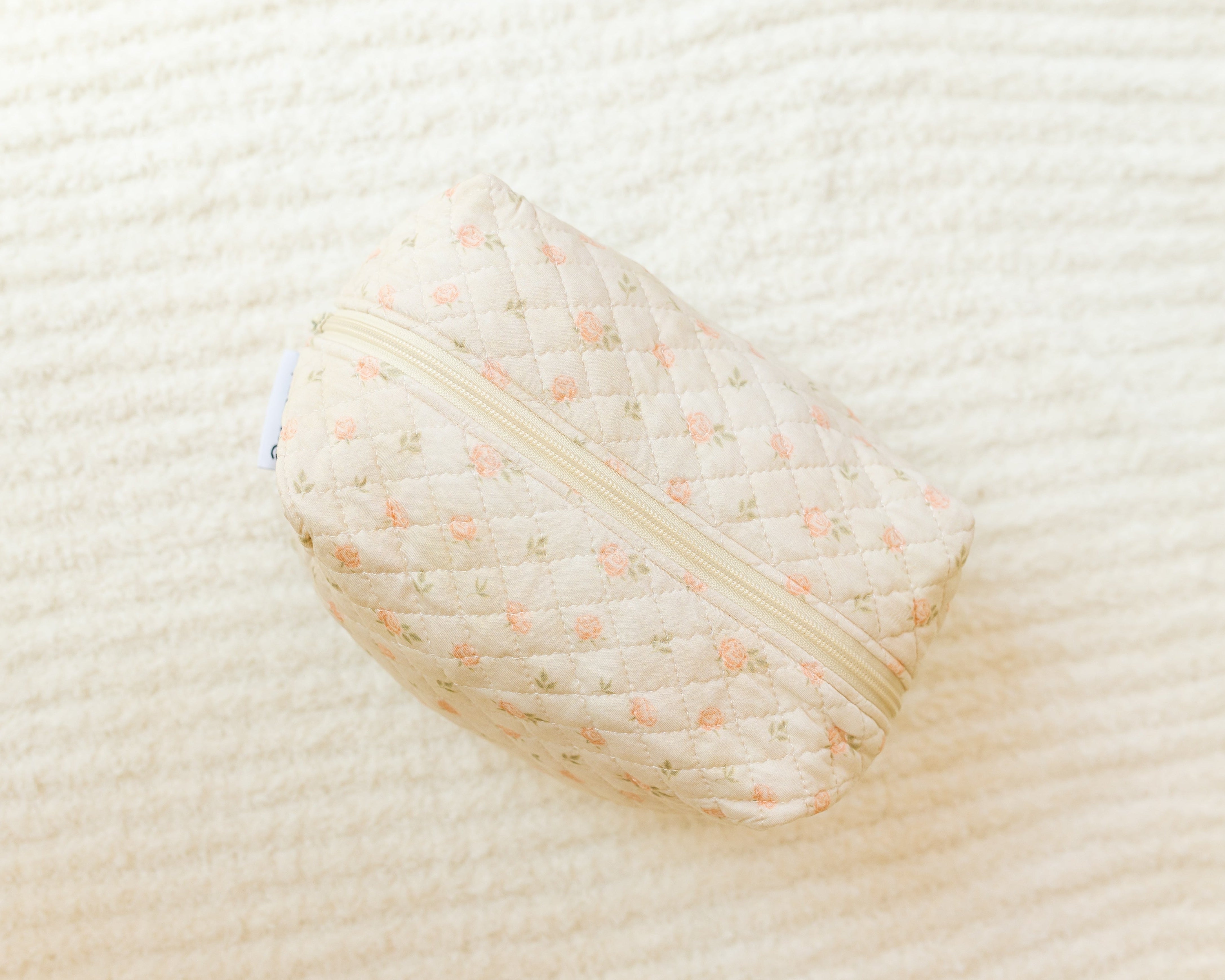 The petal / travel bag is a quilted, cream-colored travel accessory with a light floral pattern and beige zipper, shown on an off-white textured surface—ideal for organizing essentials on the go.