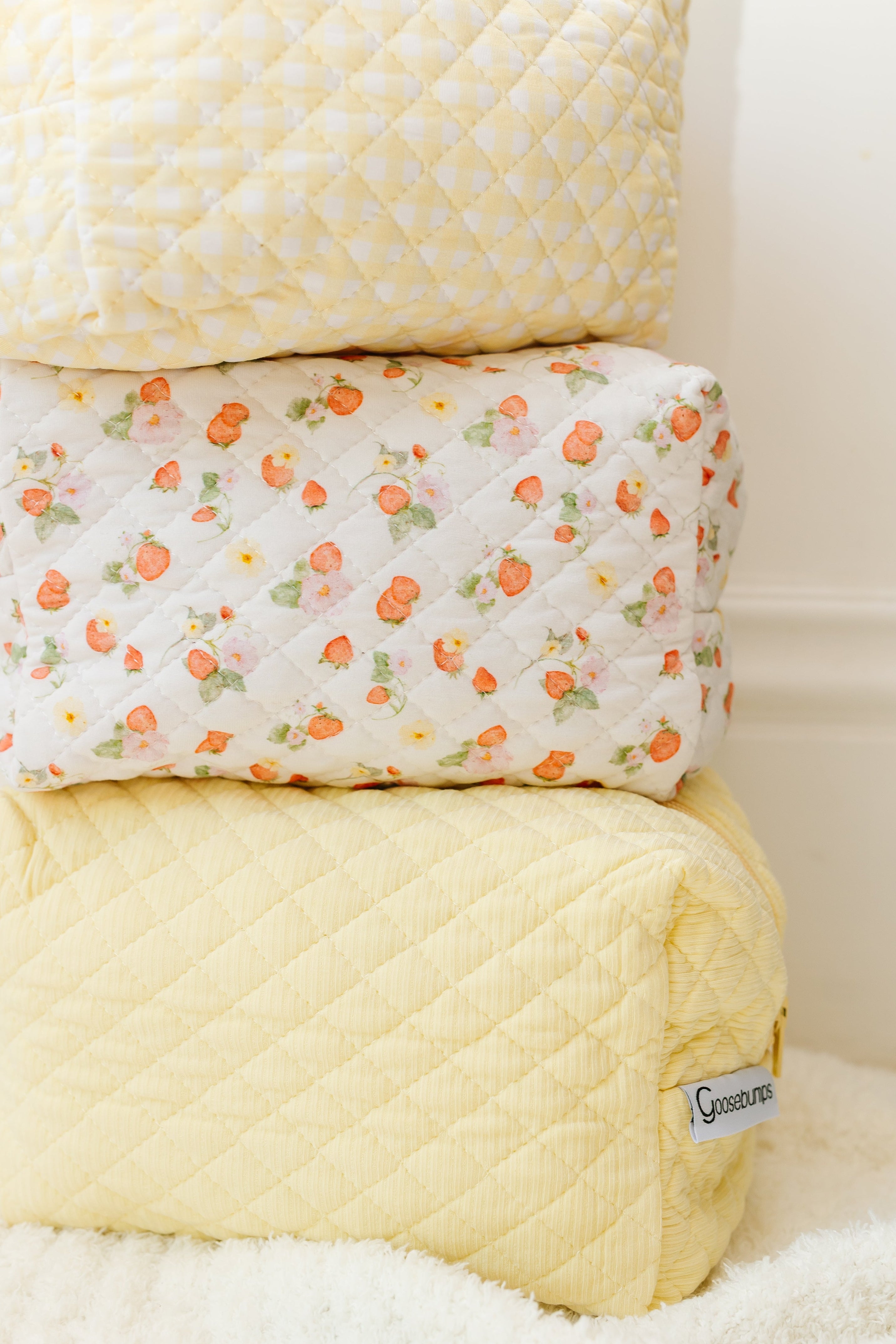 Three quilted fabric pouches are stacked: two pale yellow with diamond stitching on the top and bottom, and a strawberry-patterned pouch in the middle. The bottom freshly picked / travel bag shows a visible Gosseditions label.