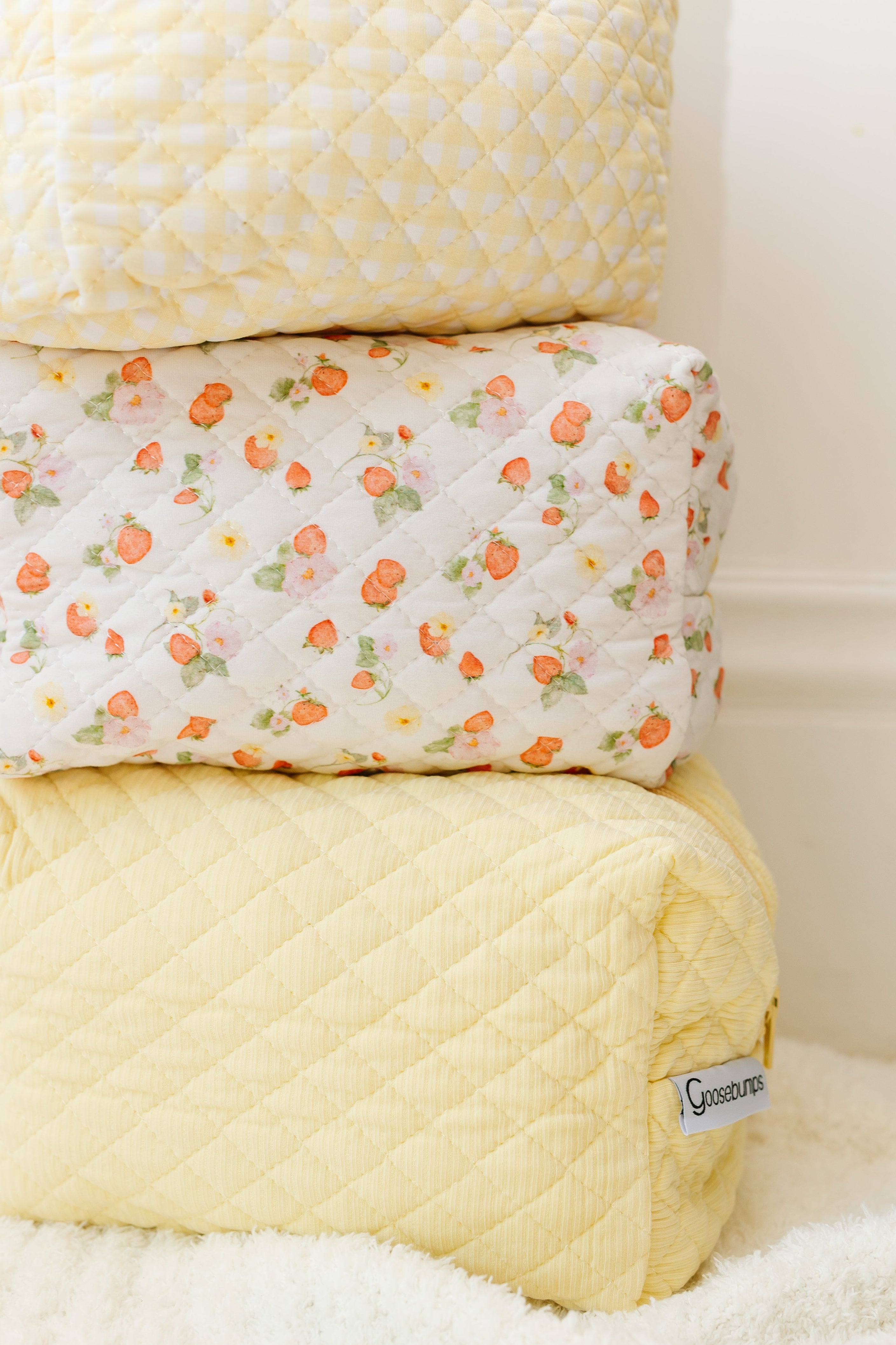 Three butter travel bags are stacked, with the top and bottom in pale yellow and the middle featuring white fabric with red strawberry and flower designs. Ideal for organizing travel essentials, they rest on a soft white surface.