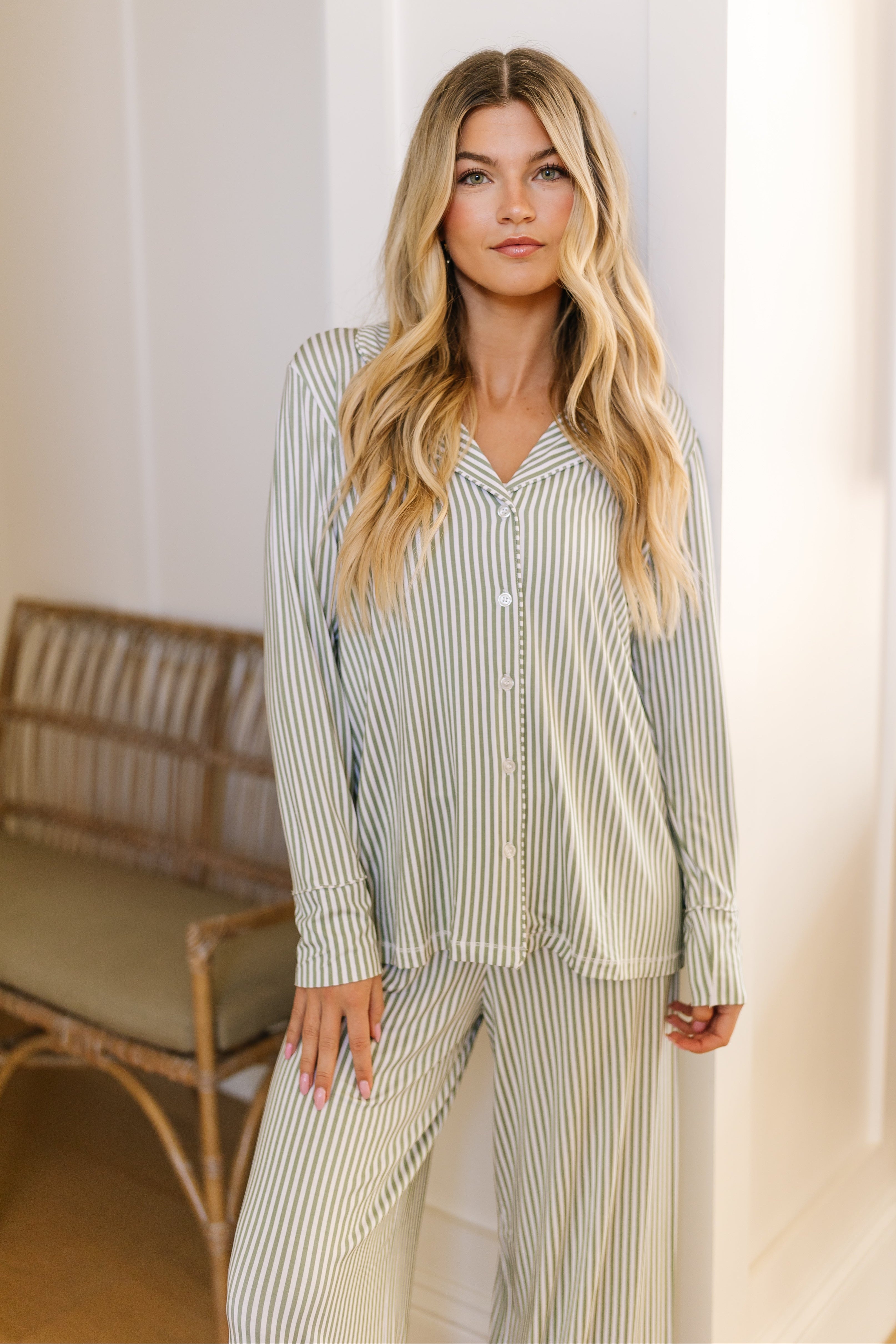 A woman with long, wavy blonde hair wears the tree house pajama set / long pant indoors. She stands by a white wall with a neutral expression, as a wooden chair and table appear in the background.
