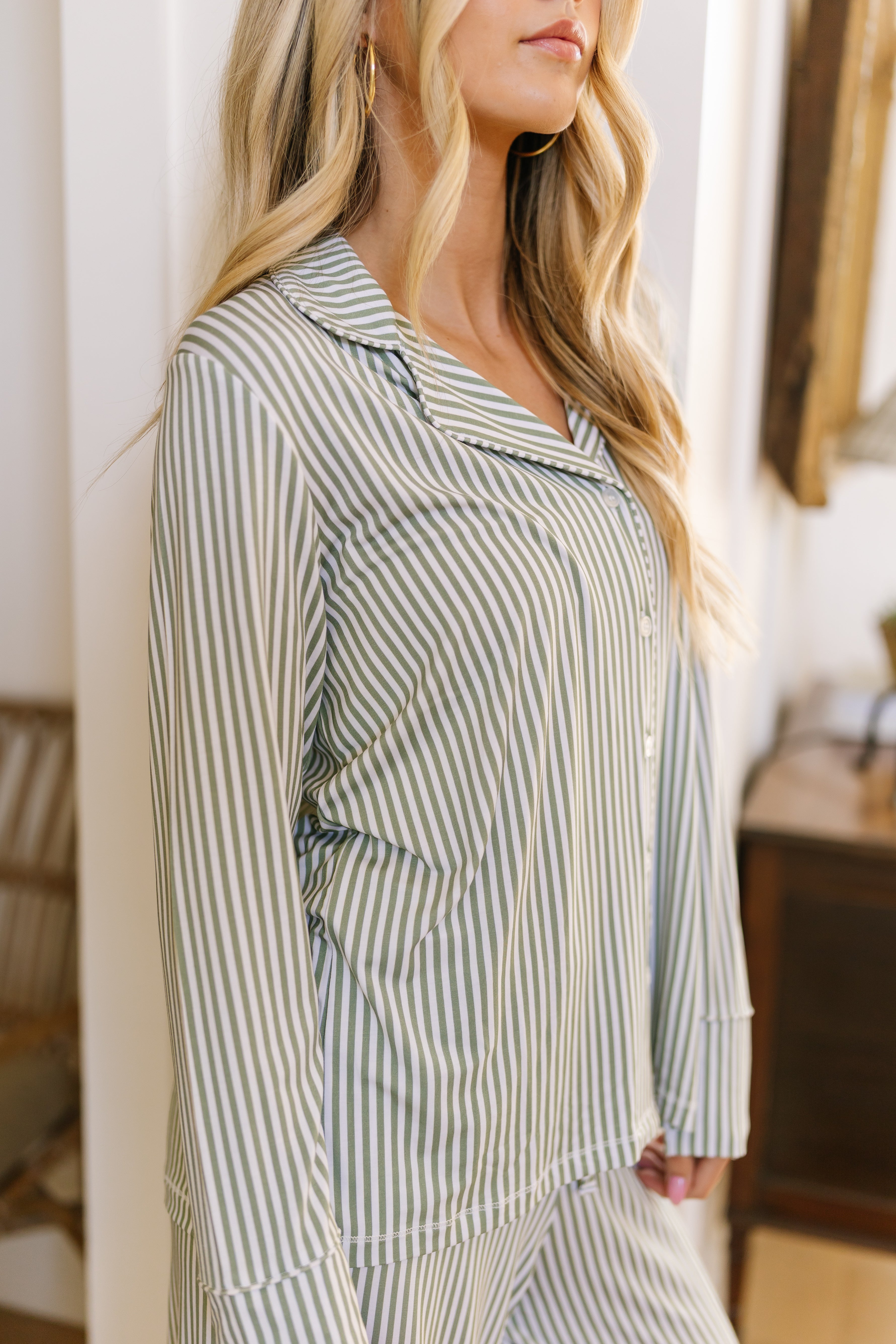 A woman with long, wavy blonde hair wears the tree house pajama set with long pants in light green and white stripes, standing indoors by a wooden chair and dresser. Her face is partially out of frame.