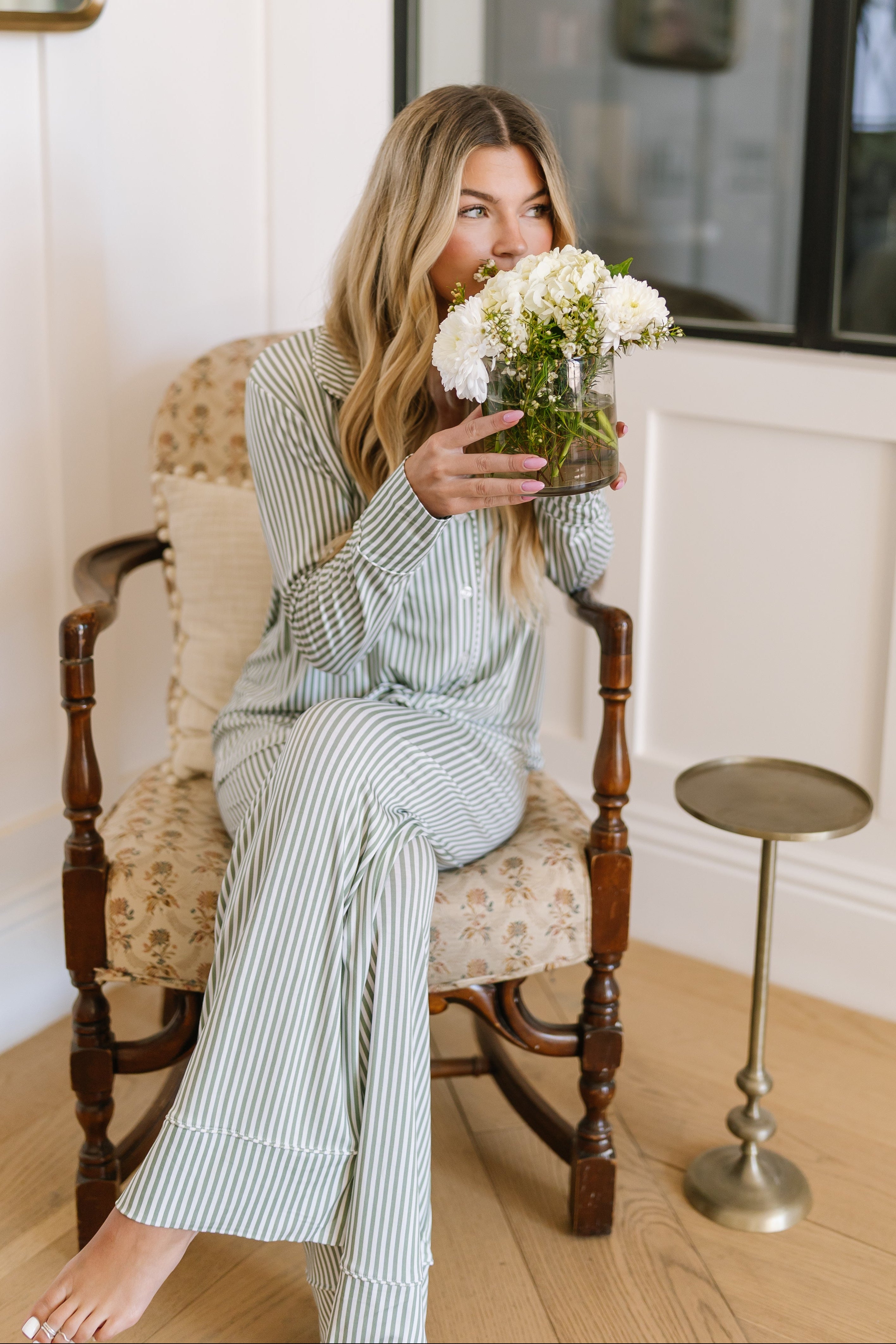 A woman wearing the tree house pajama set with long pants sits barefoot and smiling on a vintage chair indoors, holding a vase of white flowers near her face. A small round side table is beside her.
