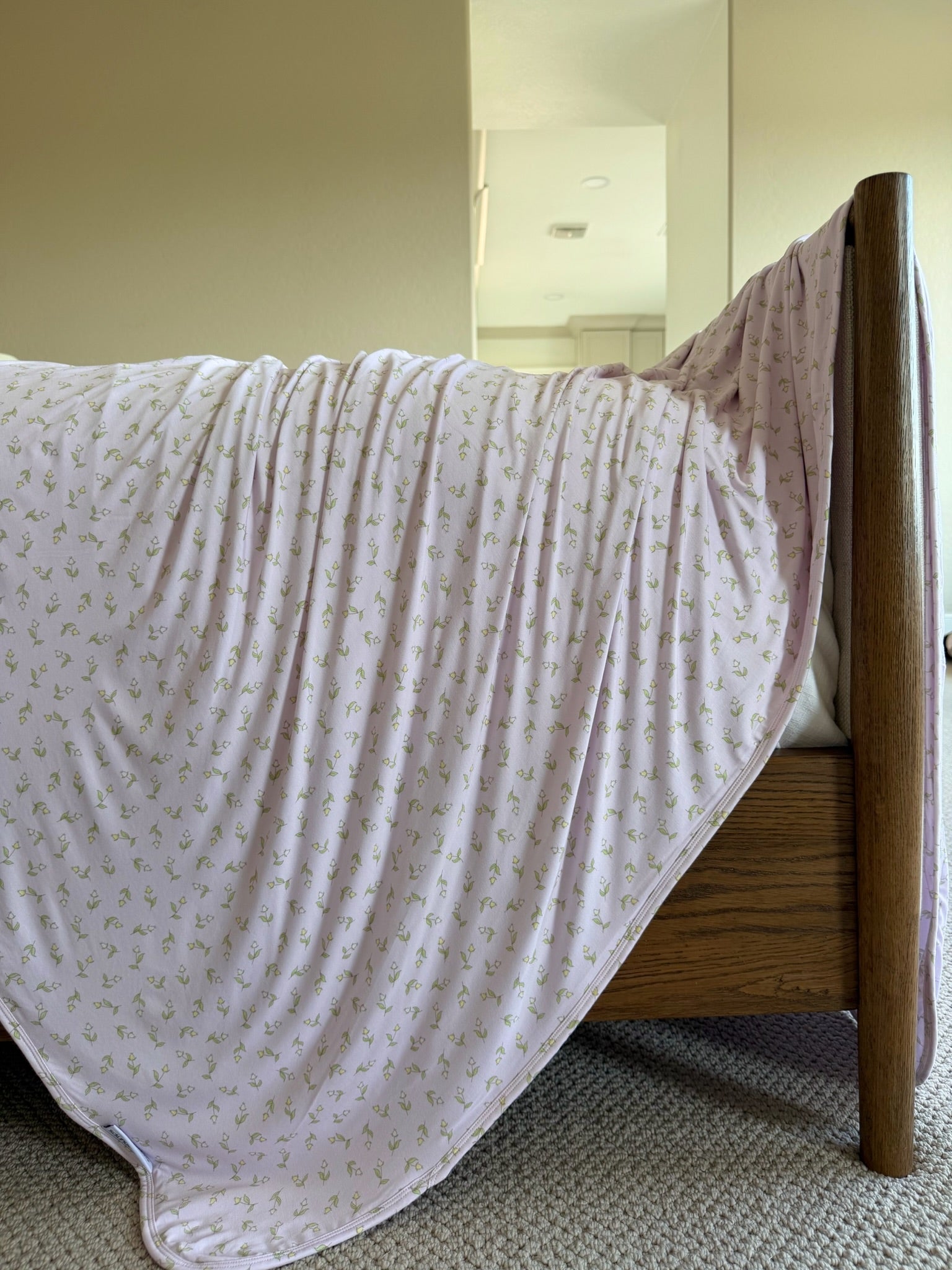 The rosebud, a light pink luxurious blanket with a subtle floral pattern, is draped over a wooden bed frame like a tent. The room features beige walls and carpeted flooring.