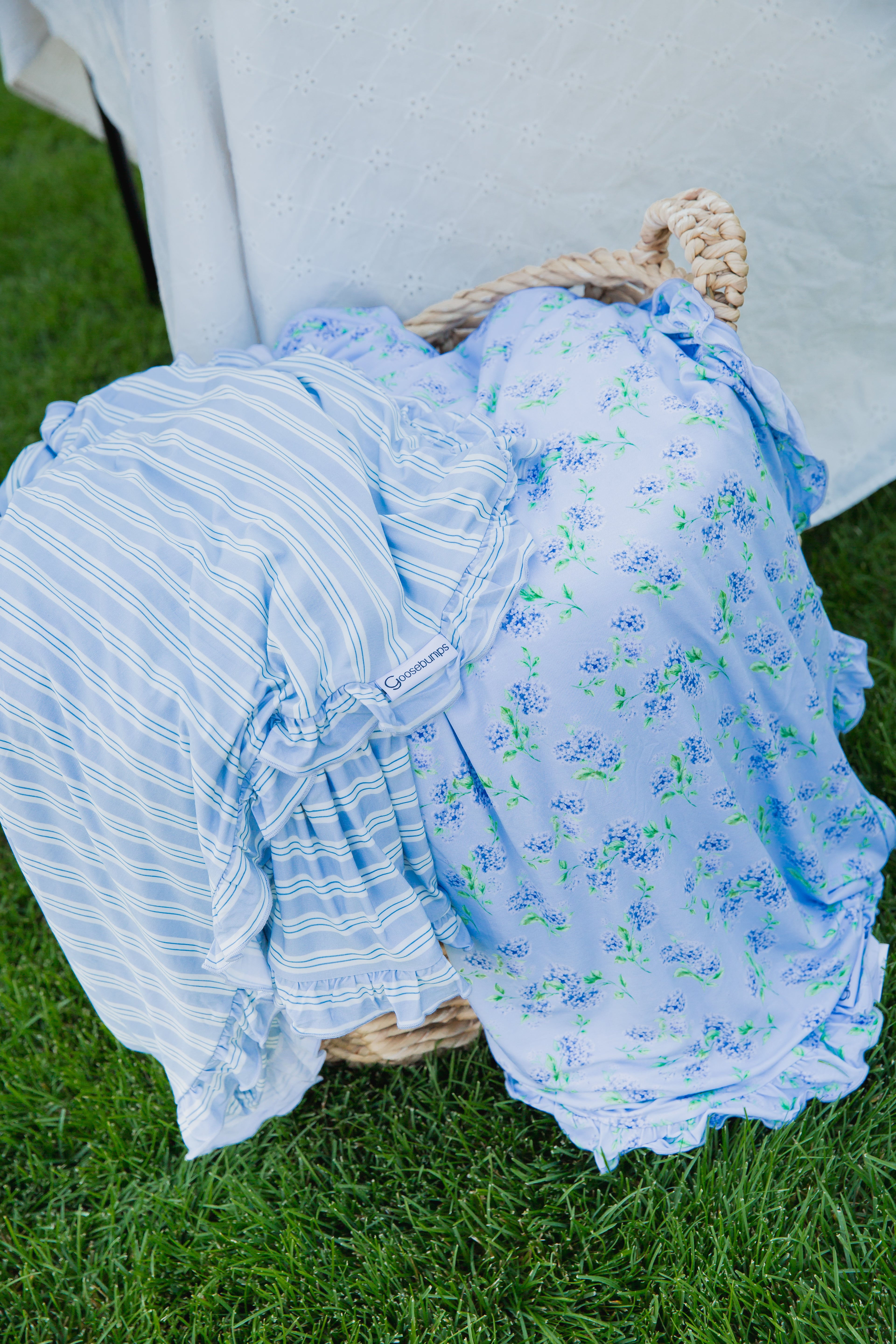 A woven basket holds two pastel dresses—one featuring blue stripes and the other showcasing the hydrangea / ruffle print—resting on green grass near a white eyelet fabric backdrop.