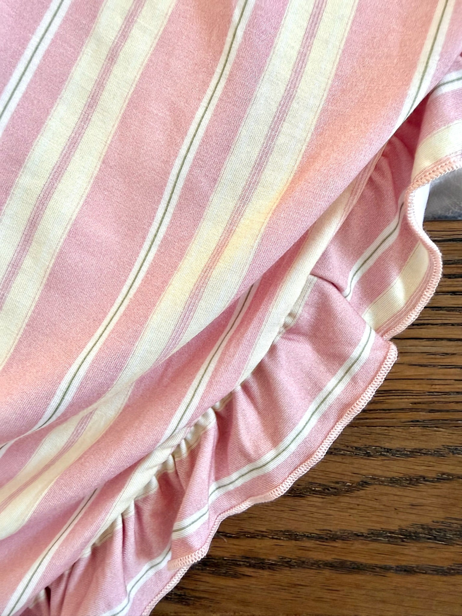 A close-up of the sherbet / ruffle bamboo blanket in pink with white and beige stripes and a ruffled edge, displayed on a brown wooden surface with visible grain patterns.
