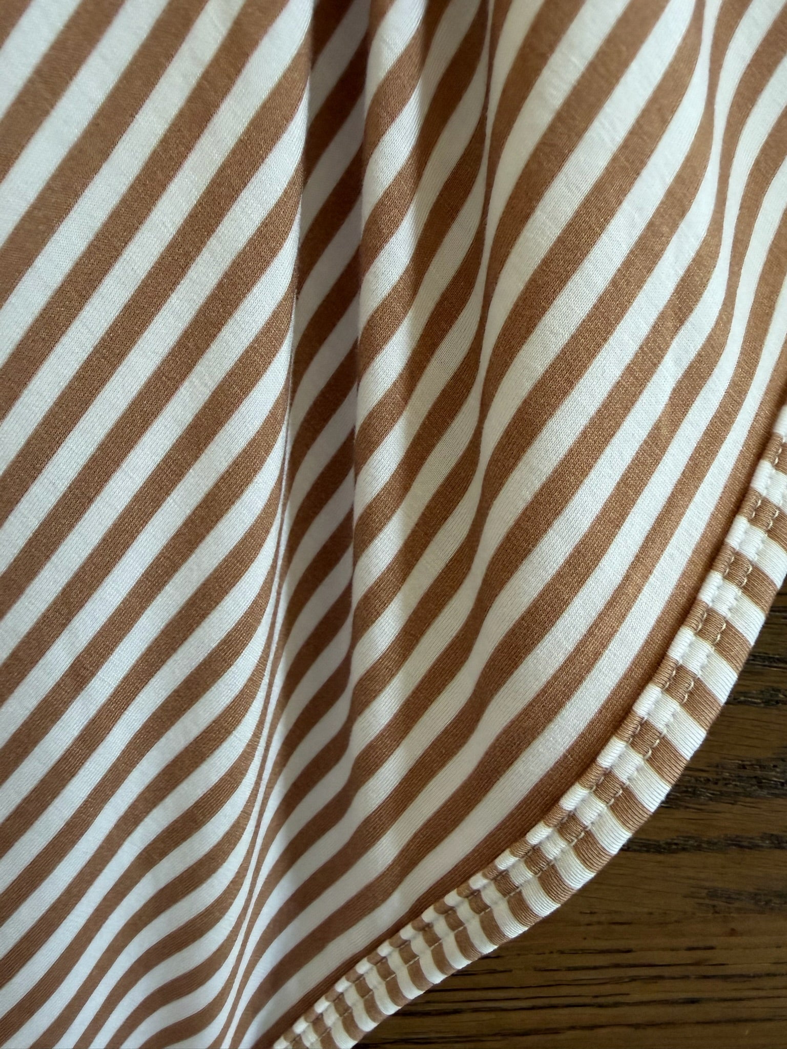 Close-up of "drift" from the Lisa Wilson Goosebumps Collection. This bamboo viscose fabric features diagonal brown and white stripes and neat stitching, offering a soft touch ideal for an adult swaddle. Displayed resting on wood.