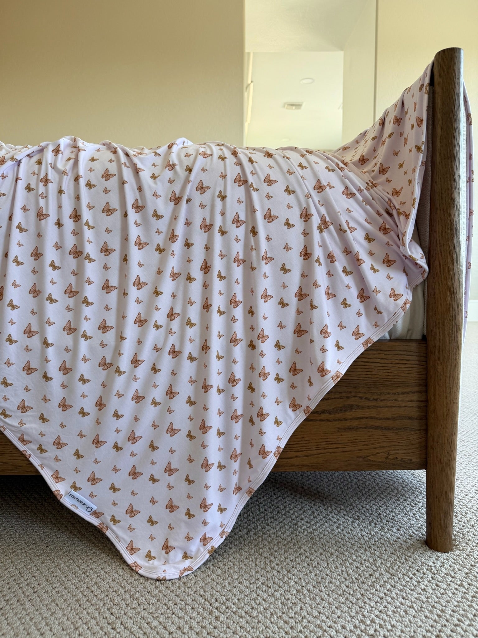 A light pink "flutter" bamboo blanket with a butterfly print hangs over a wooden bed frame in a sunlit, carpeted room.