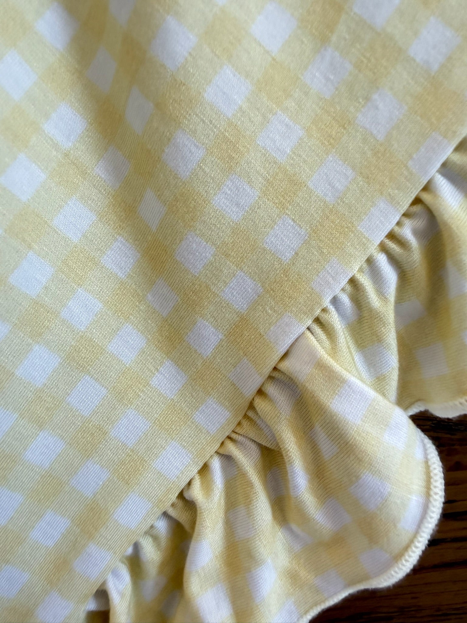 The "sunshine / ruffle" blanket features yellow and white gingham with a delicate ruffled edge and small white squares on a pale yellow background, adding charming detail to this luxurious piece, shown close up on a wooden surface.