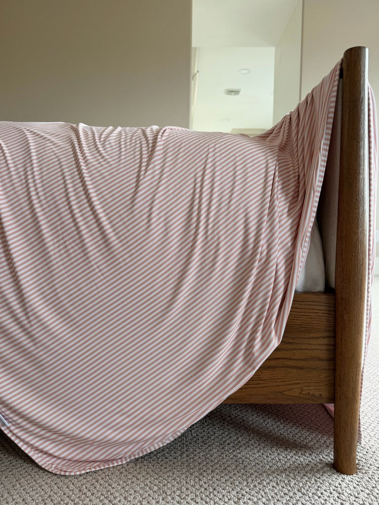 A luxurious Heritage blanket with pink and white stripes drapes over the side of a wooden bed frame in a carpeted room with neutral-colored walls.