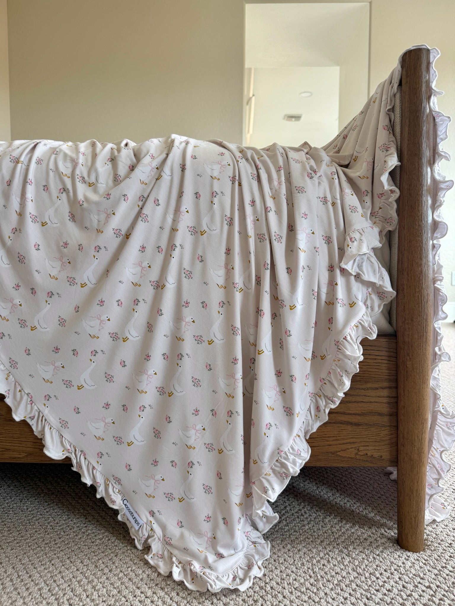 The mother goose / ruffle blanket, featuring a floral print and ruffled edges, drapes over a wooden bed frame and touches a beige carpeted floor in a softly lit room.