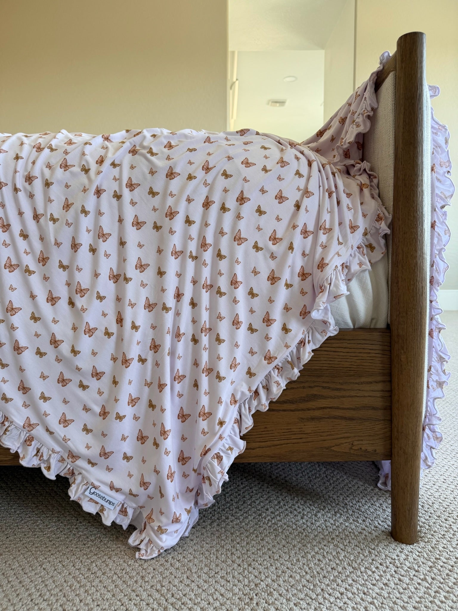 The flutter/ruffle pink butterfly-patterned blanket with delicate ruffle trim is draped over a wooden bed, set on a beige carpet in a bright, neutral room.