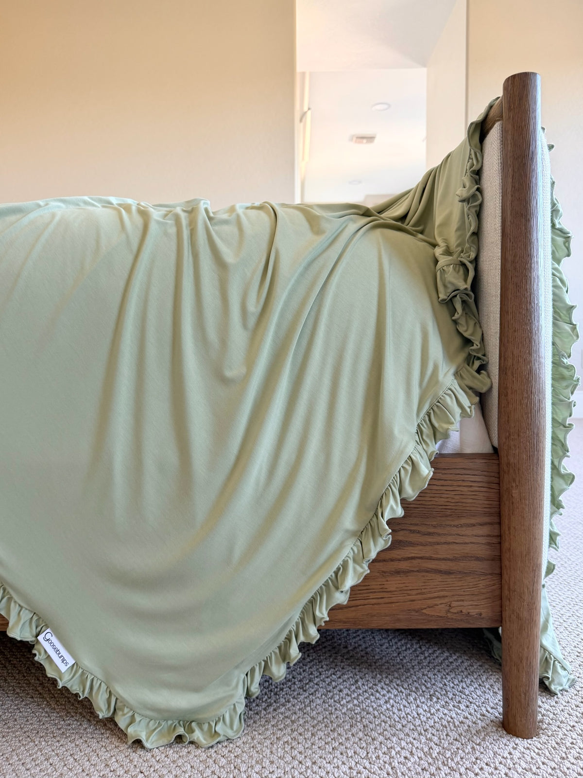 Frosted fir / ruffle sage green bamboo blanket with a ruffled edge draped over the corner of a wooden bed frame.