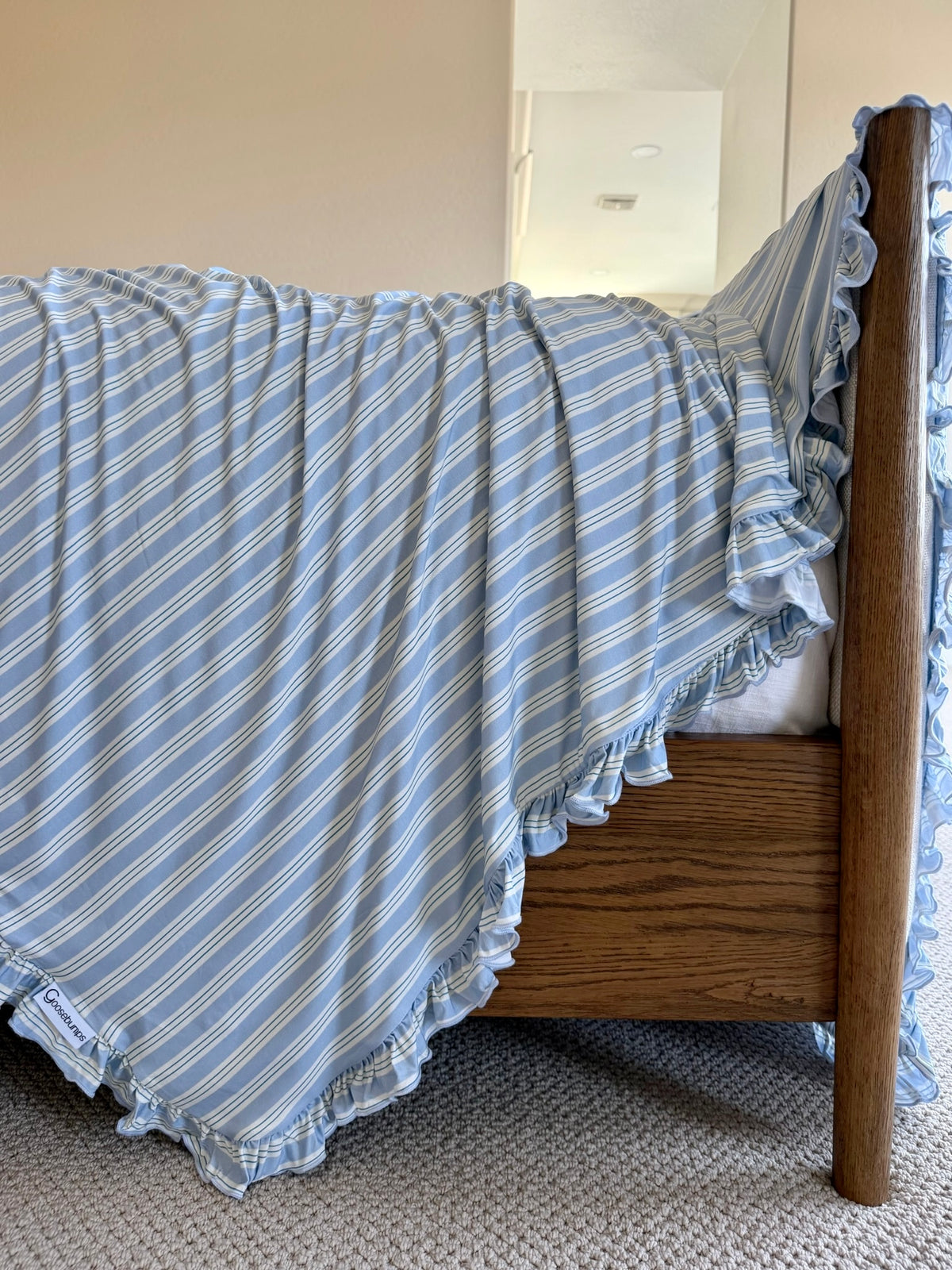 A haven / ruffle blanket in blue and white stripes is draped casually over the side of a wooden bed frame in a carpeted room with neutral walls.