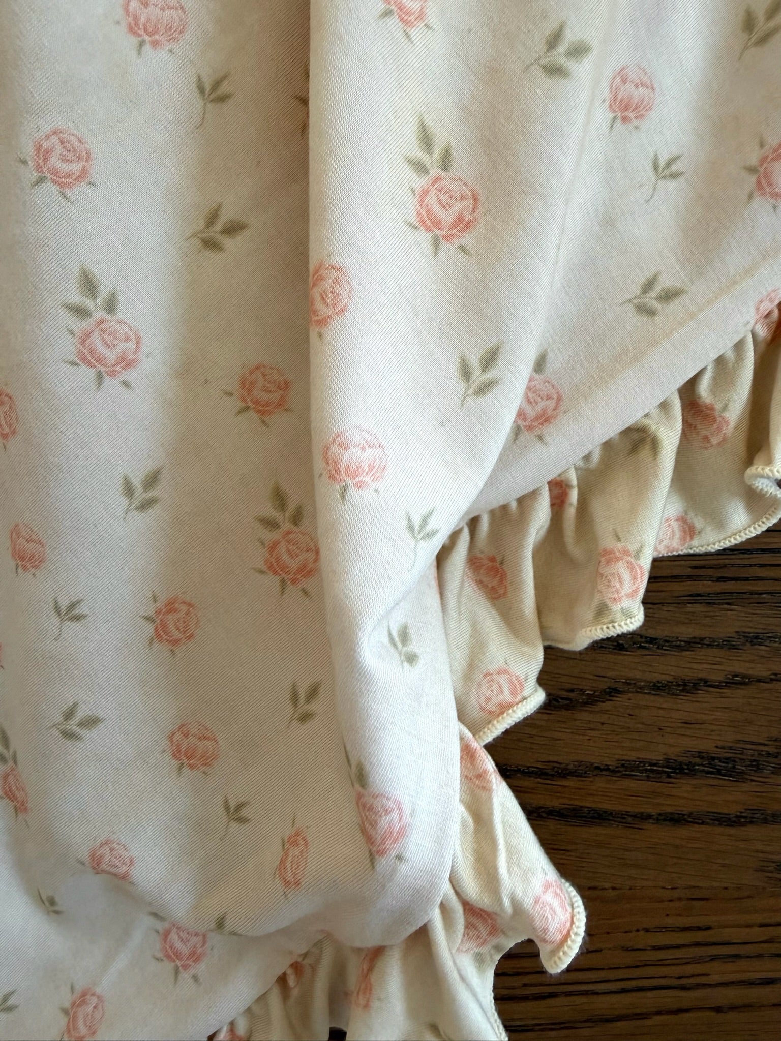 The petal / ruffle blanket, cream-colored with small pink rose and green leaf patterns plus ruffled edges, is draped over a wooden surface.