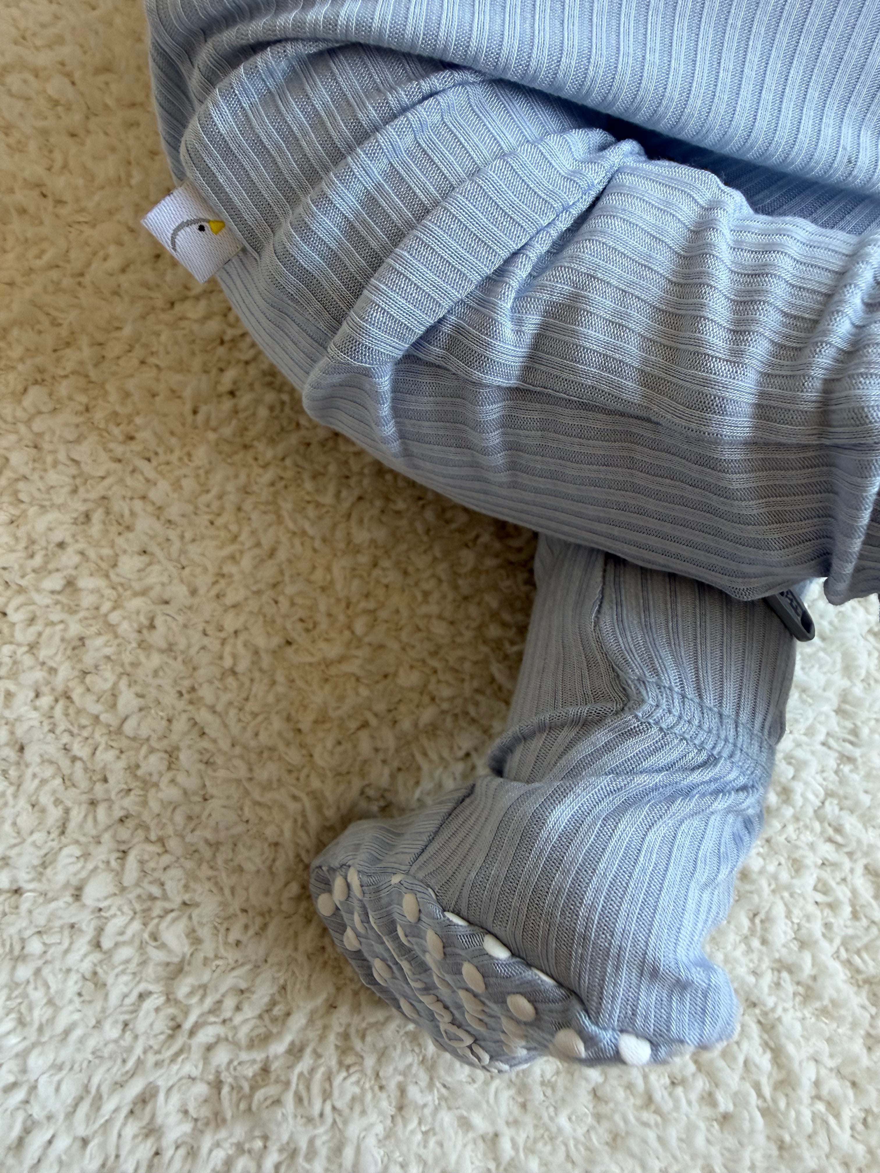 A baby wearing the cove one piece zipper sits on a soft cream carpet. The baby’s feet, covered by the footed pajamas with white grippers, are visible and crossed at the ankles.