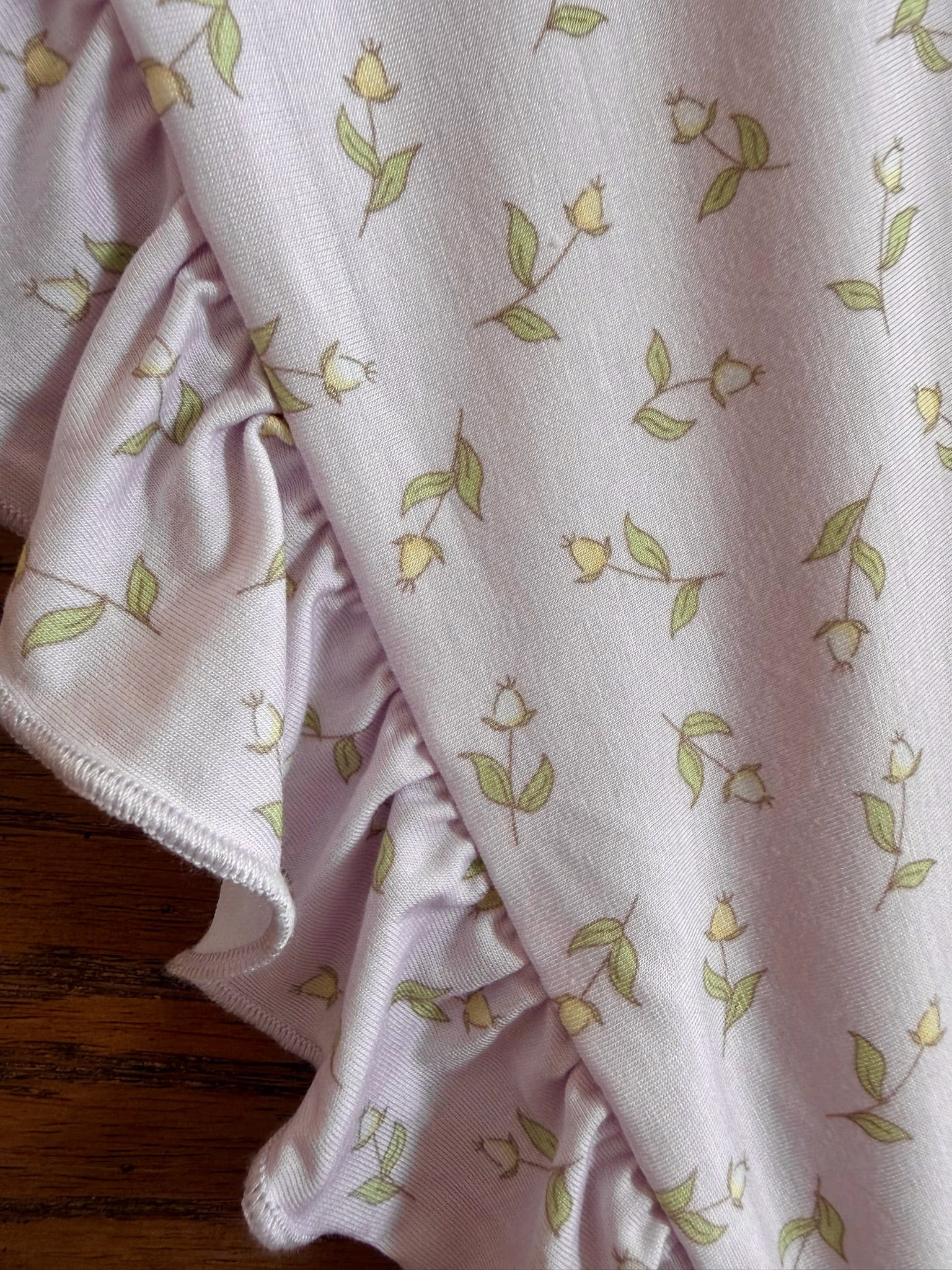The rosebud / ruffle blanket features light purple fabric with small yellow flower buds and green leaves, finished with ruffled trim on one edge and displayed on a wooden surface.