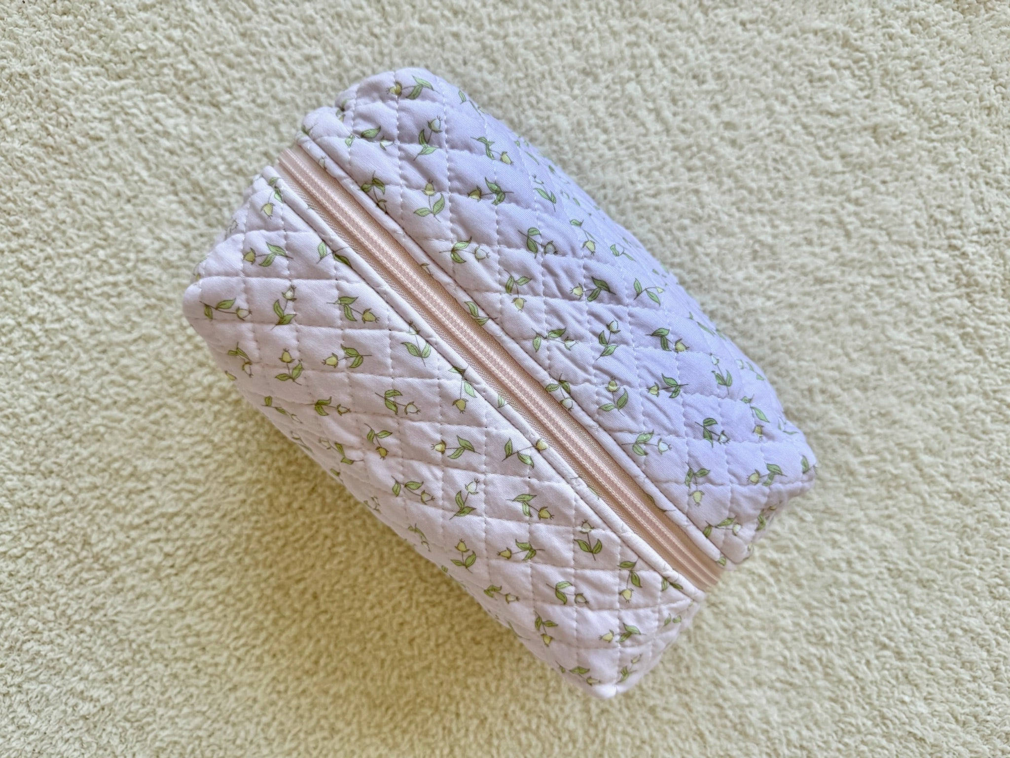 A quilted, rectangular pink pouch adorned with delicate green floral patterns and finished with a light pink zipper sits atop a textured cream surface—perfect as the stylish rosebud / travel bag for carrying your favorite travel essentials.